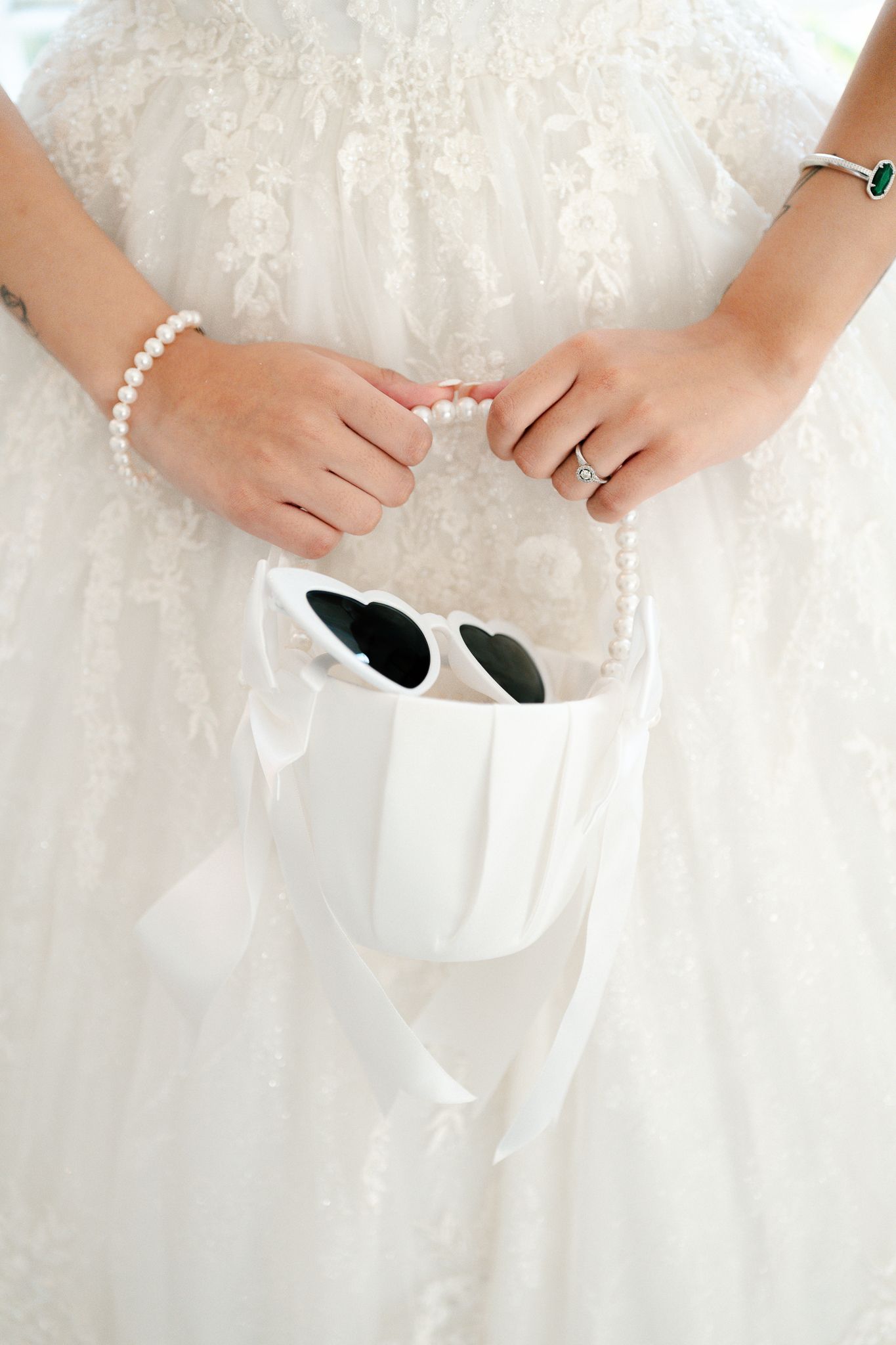 Bride in white lace gown holding pearl necklace and white handbag with heart-shaped sunglasses, wearing pearl bracelet and ring.