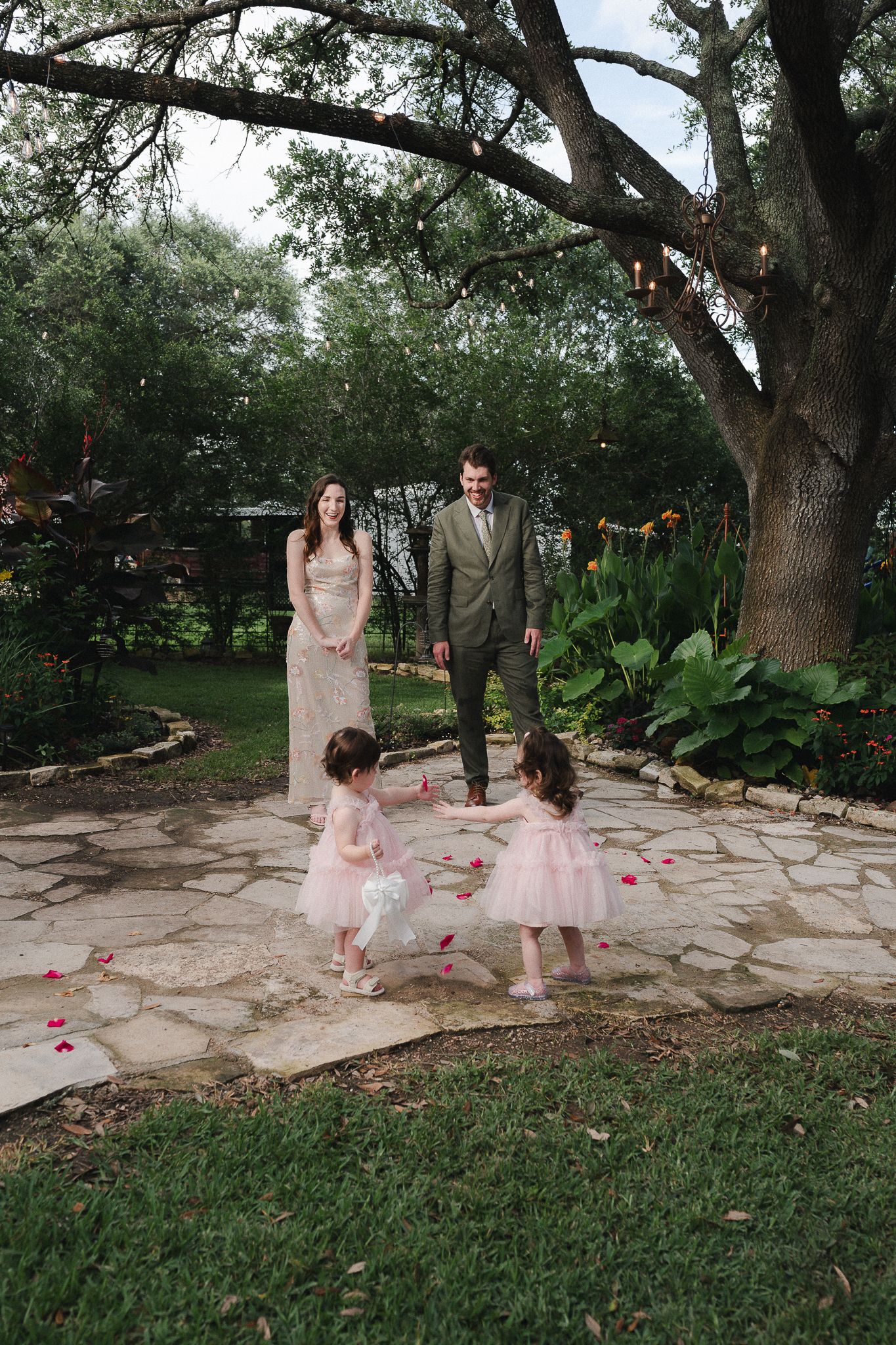Bride and groom watch two flower girls in pink dresses scatter rose petals on stone path in lush garden wedding ceremony.