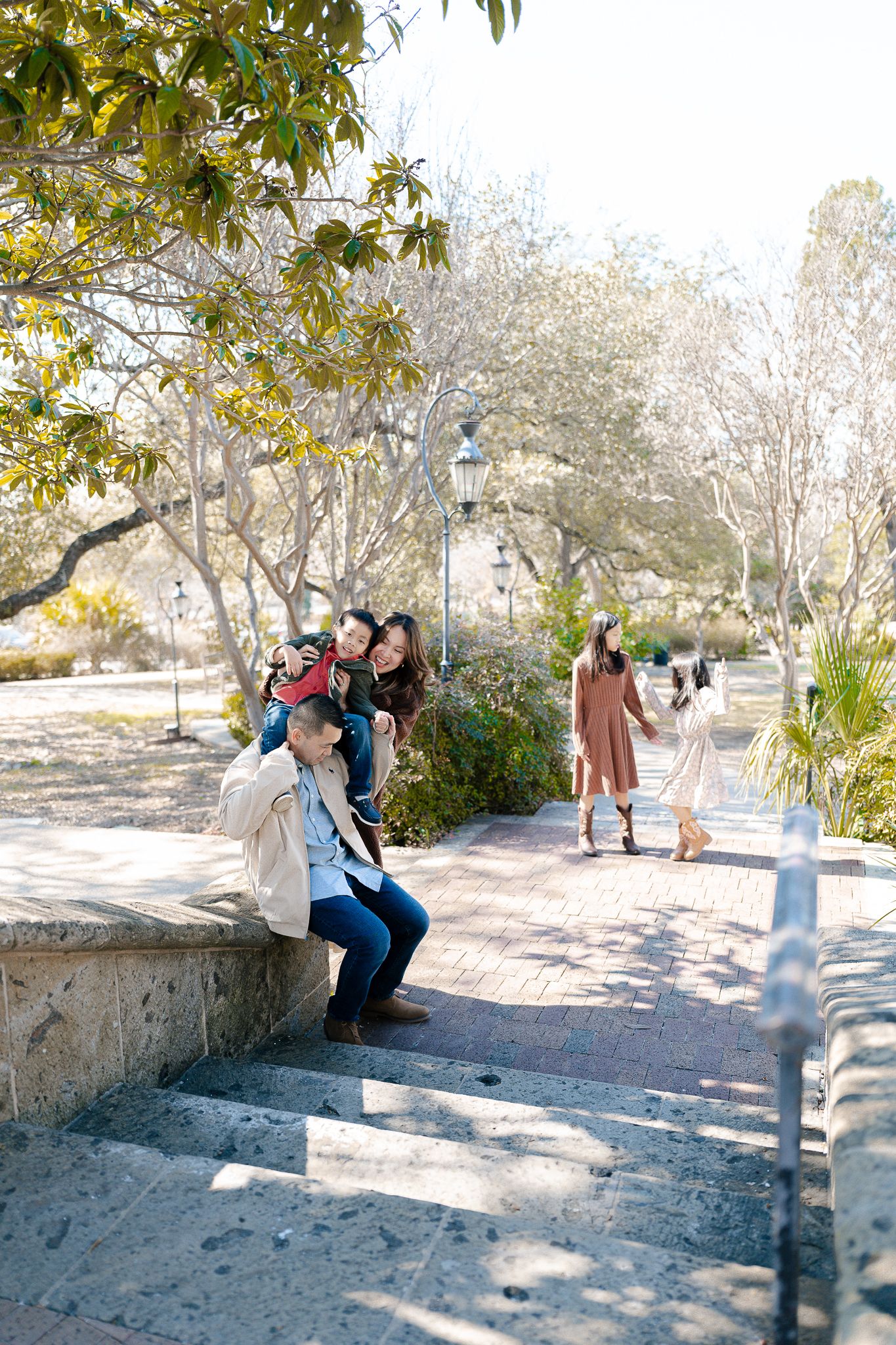 People enjoying a sunny day in a park with stone steps, trees and lamp posts along a walkway.