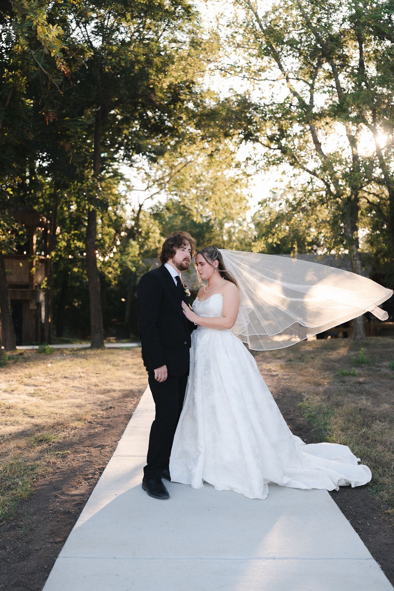 Bride and groom share a romantic moment on a pathway, her veil flowing in the breeze, surrounded by lush trees at golden hour.