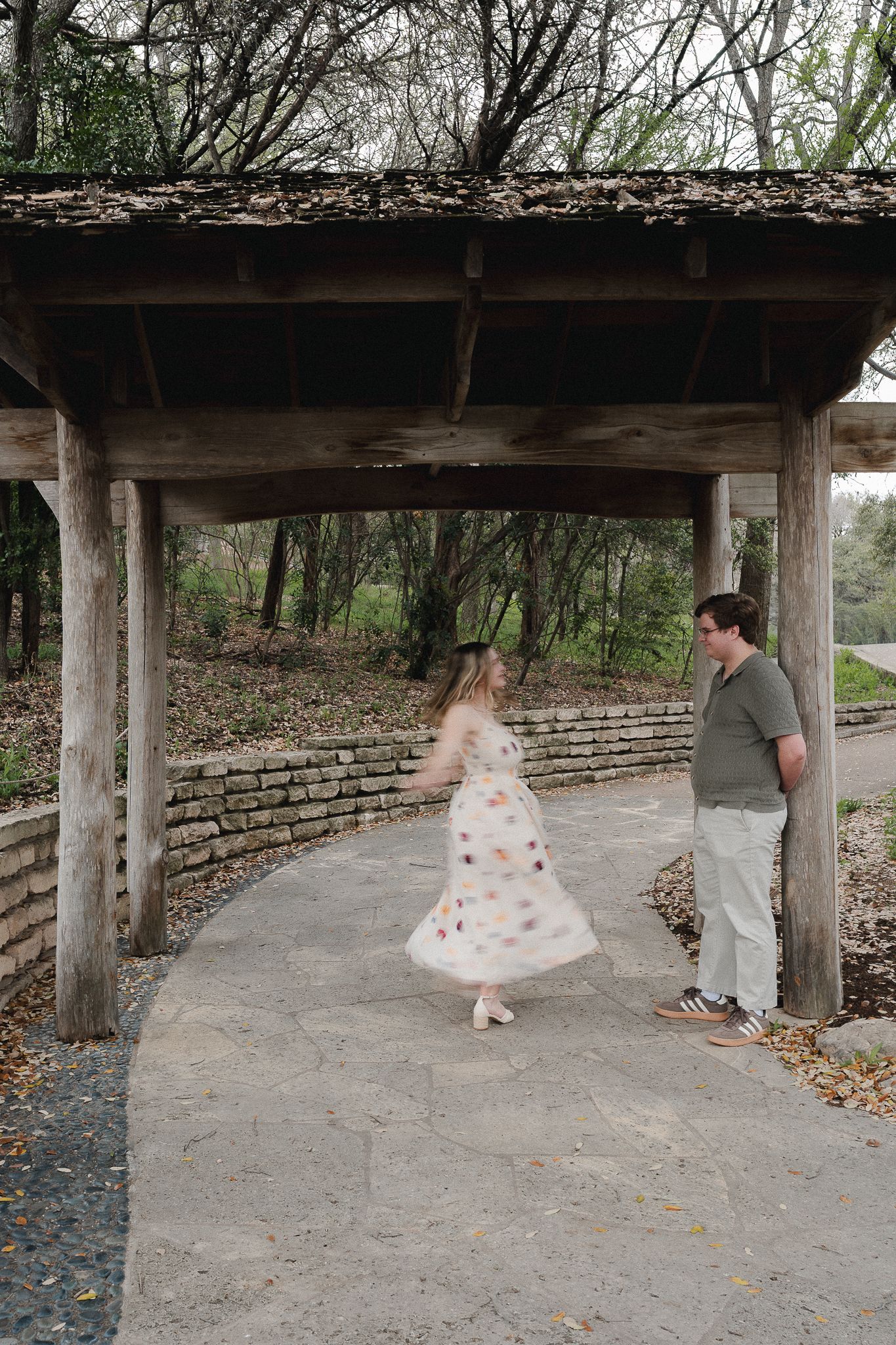 Couple sharing a romantic moment under a rustic wooden pergola in a lush park setting.