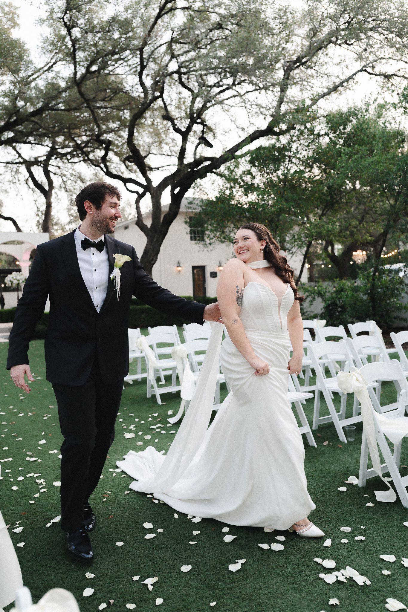 Bride and groom walk joyfully down petal-strewn aisle after outdoor wedding ceremony surrounded by white chairs.