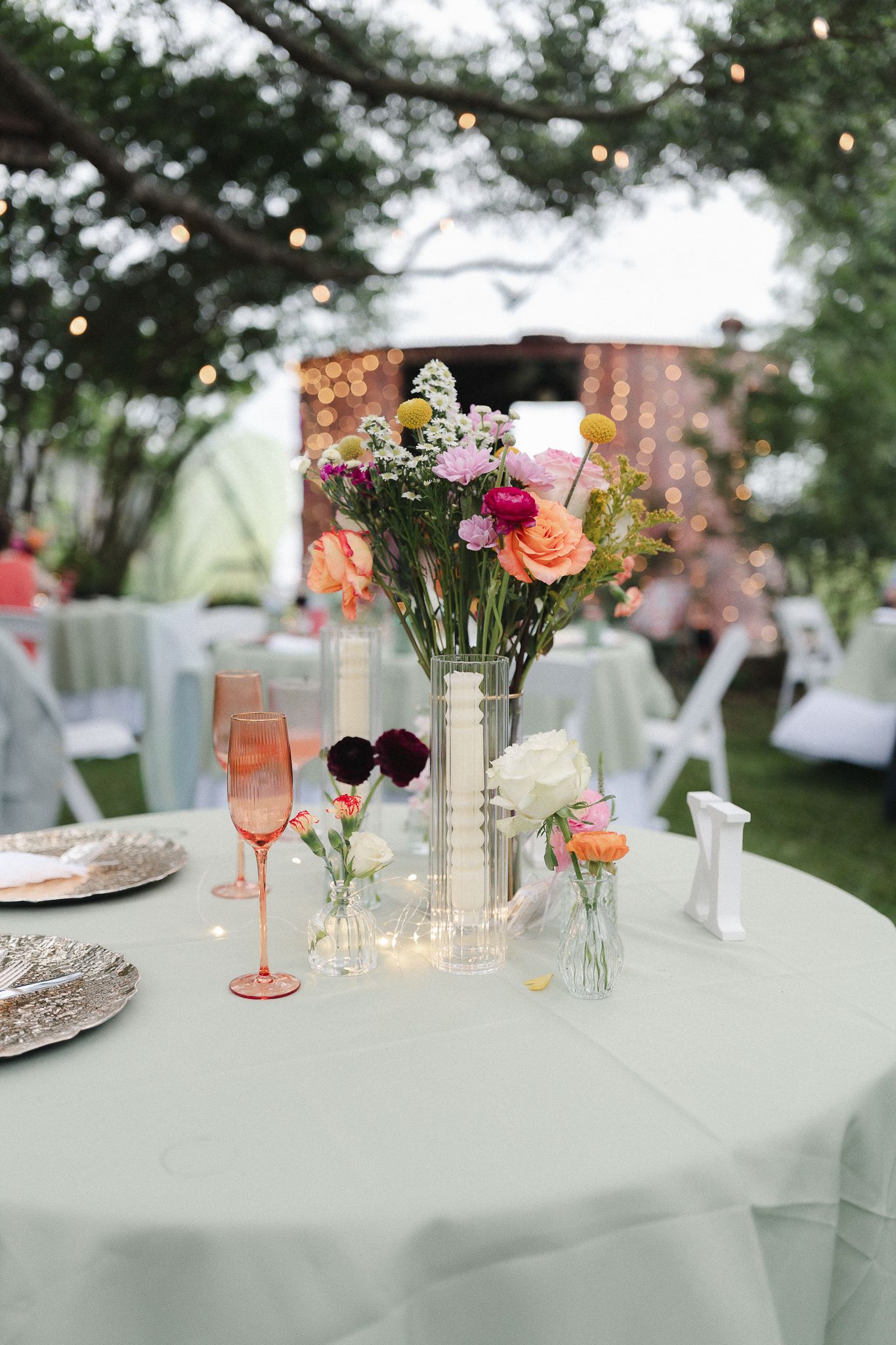 Elegant outdoor wedding reception table with colorful floral centerpiece, pink champagne glasses, and string lights in background.
