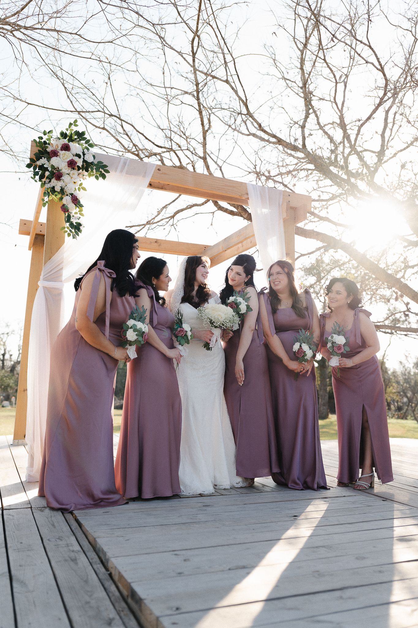 Bride in white gown poses with five bridesmaids in mauve dresses holding bouquets under a wooden arch outdoors.