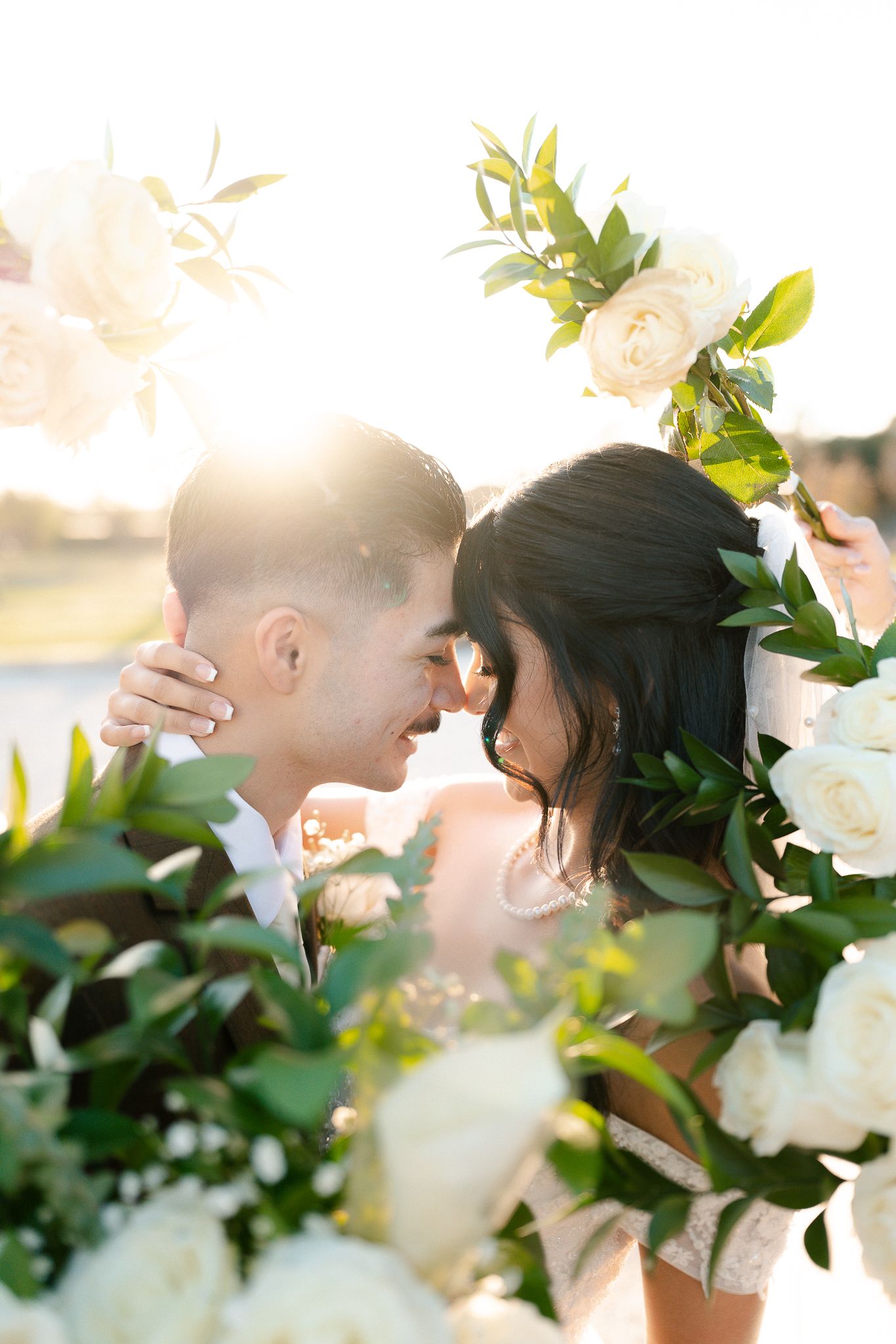 Couple sharing a romantic moment surrounded by white roses and greenery with golden sunset backlight at their wedding.