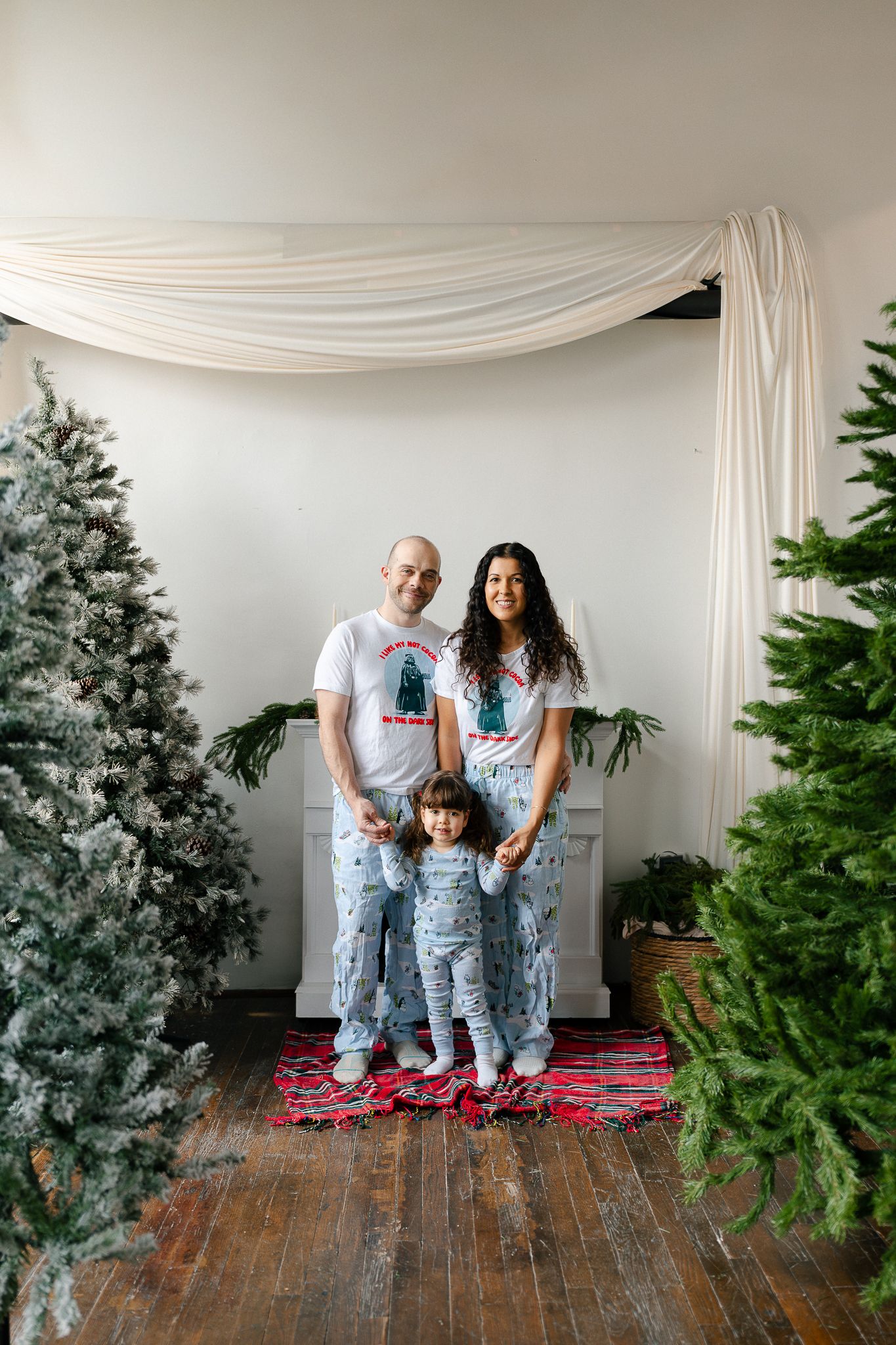 Family wearing matching blue snowflake pajamas and red slippers pose in front of Christmas trees against white backdrop.