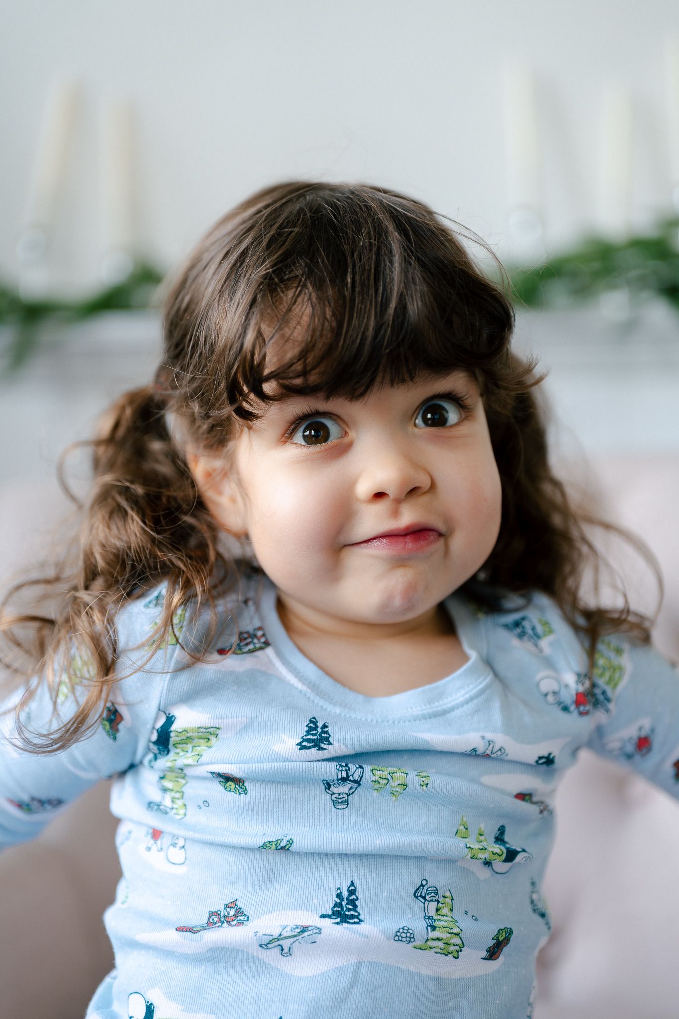 A young child with dark curly hair and bright blue eyes wearing a light blue patterned shirt makes an expressive face.