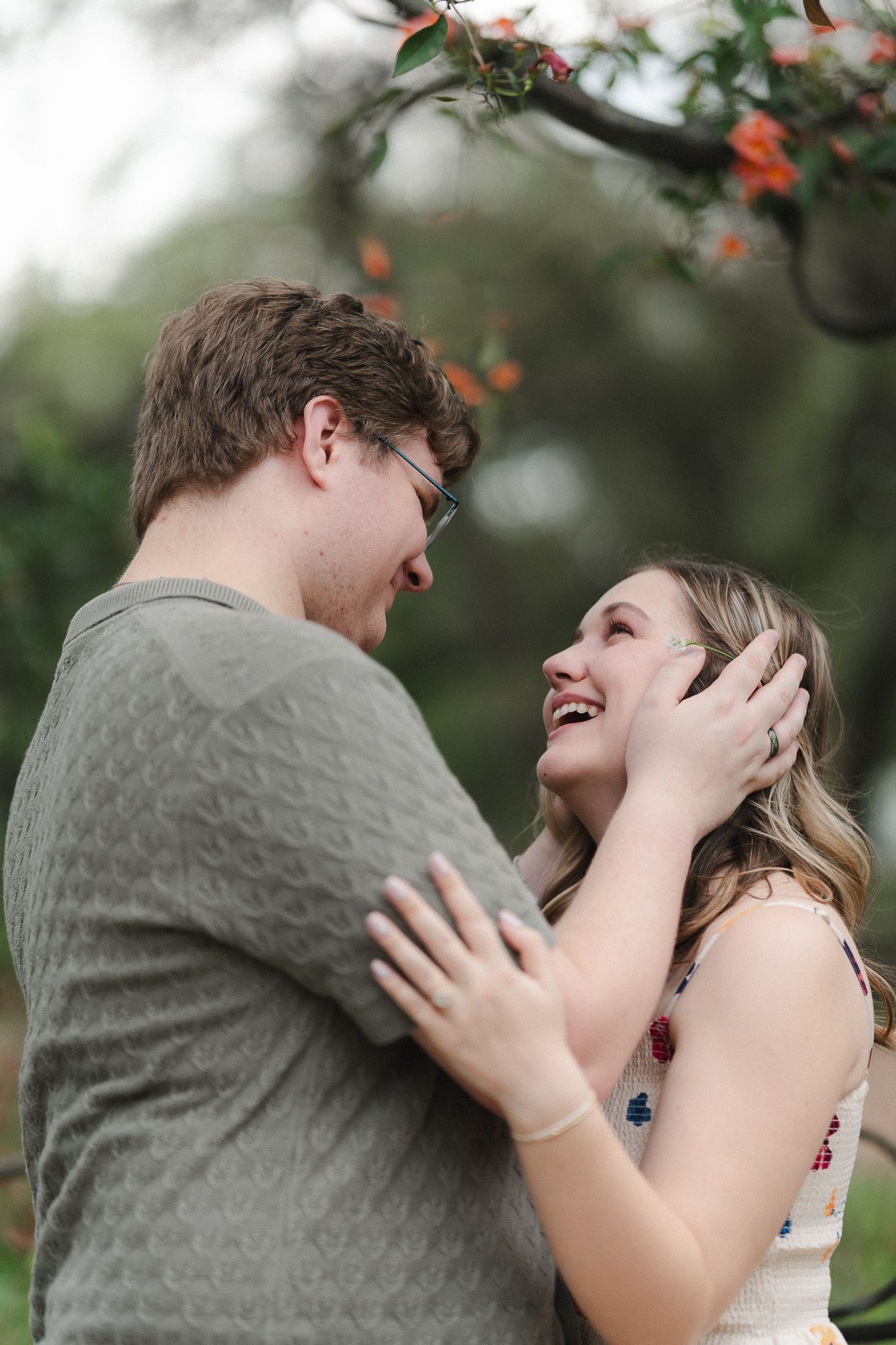 A laughing couple embraces outdoors under flowering branches, the woman in a floral dress gazing up at her partner.