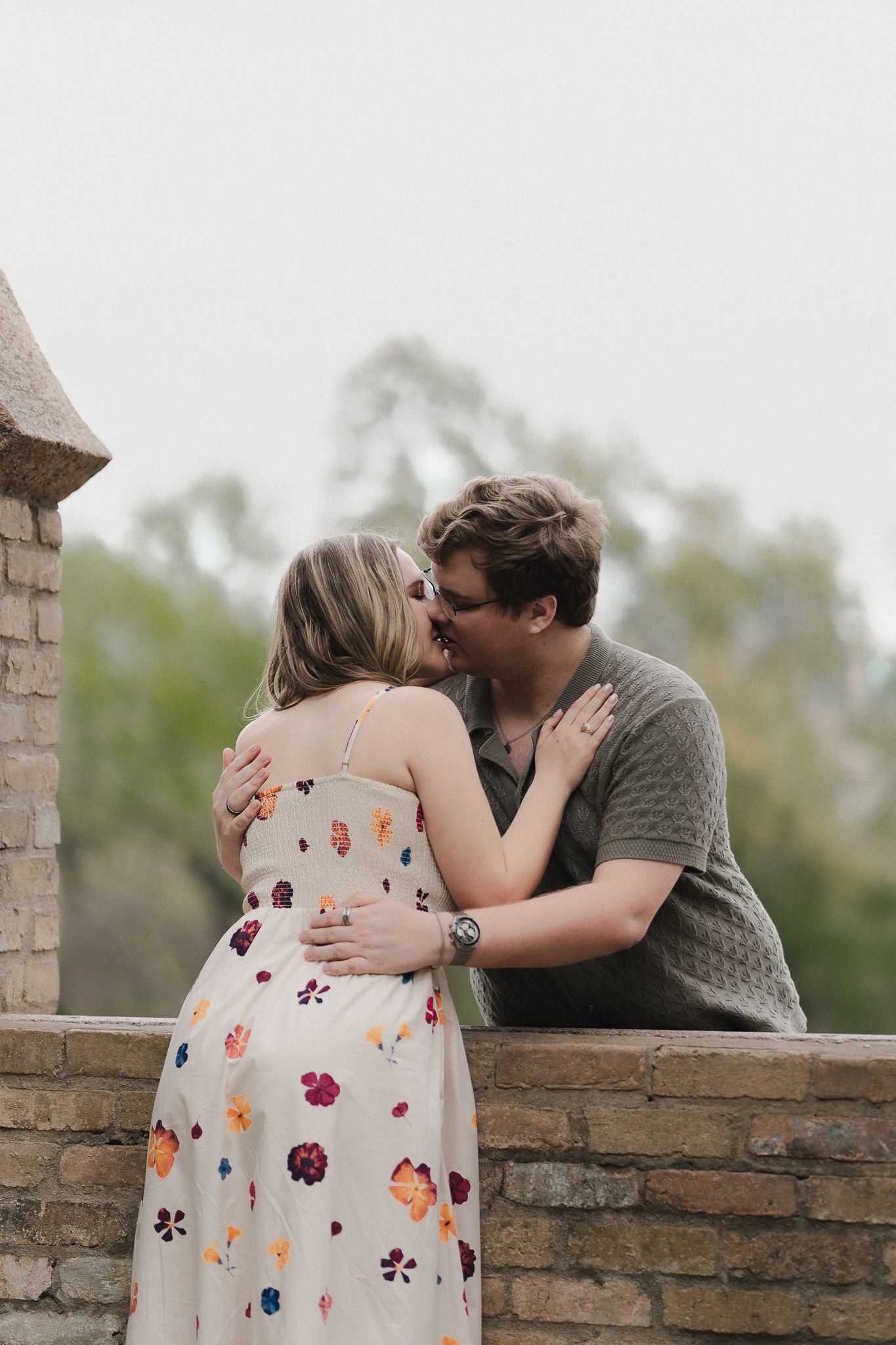 A couple shares a romantic kiss on a brick terrace, the woman wearing a floral sundress and the man in a green shirt.