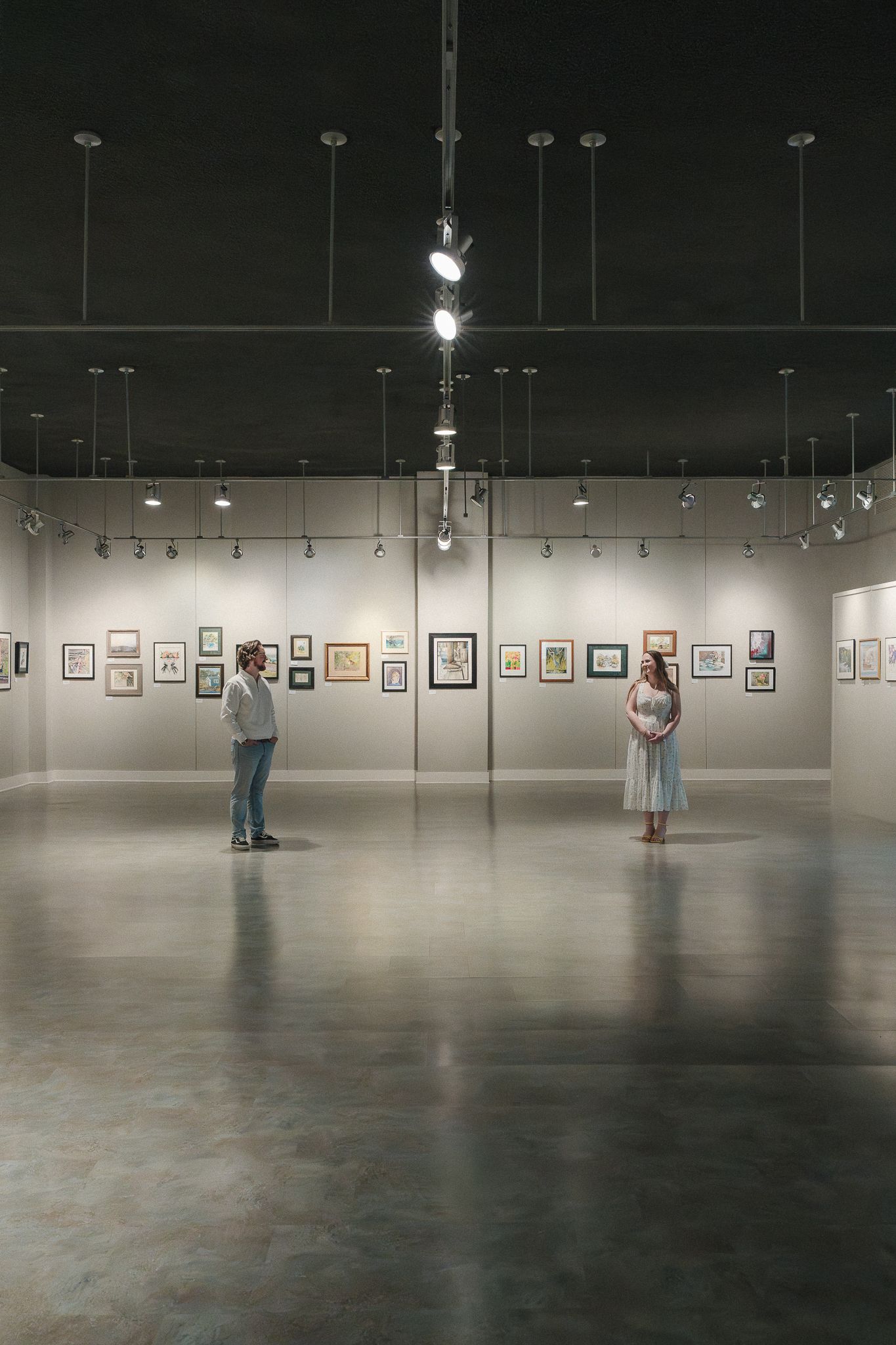 Two people standing apart in a modern art gallery with framed artwork on white walls and dramatic ceiling lighting.