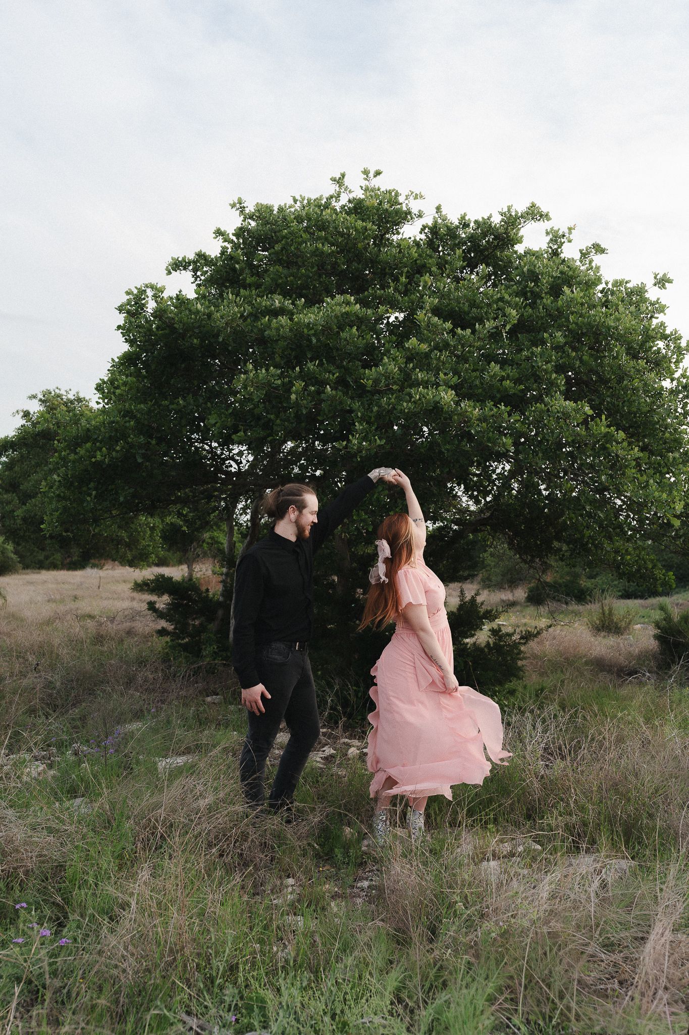 Couple dancing outdoors near a large green tree, woman in pink dress holding flowers, man in black outfit.