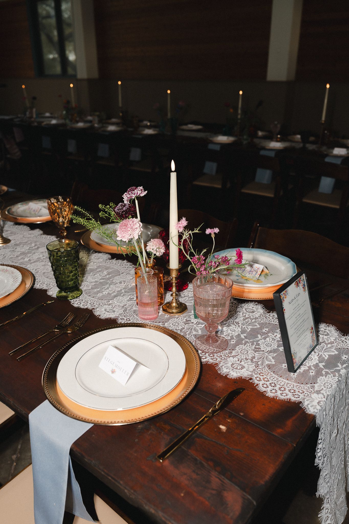 Elegant wedding reception table with lace runner, gold chargers, taper candles, and pink floral centerpieces on dark wood.