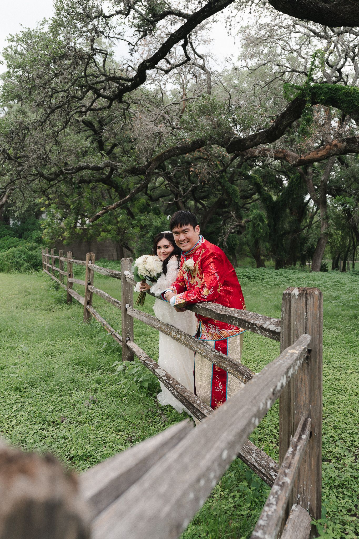 Couple in traditional Chinese wedding attire posing by a rustic wooden fence surrounded by lush green trees.