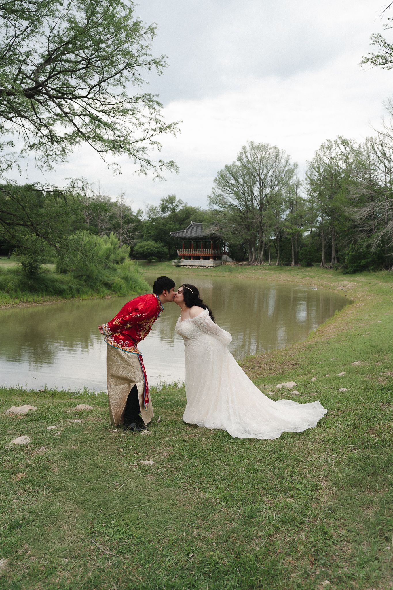 Couple in traditional wedding attire sharing a kiss by a serene pond with a pavilion backdrop.