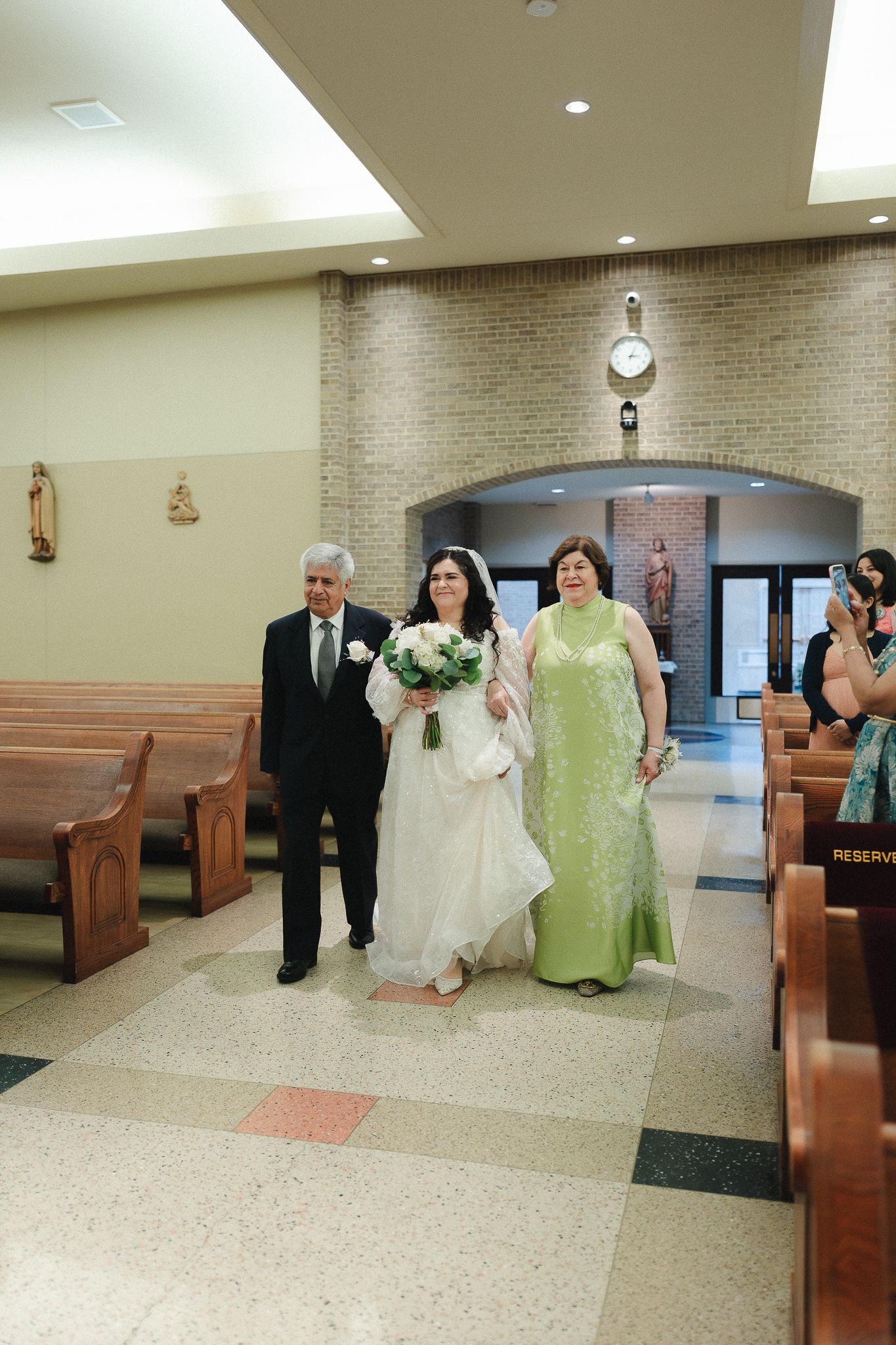 Bride walks down church aisle escorted by parents, carrying white bouquet during wedding processional.