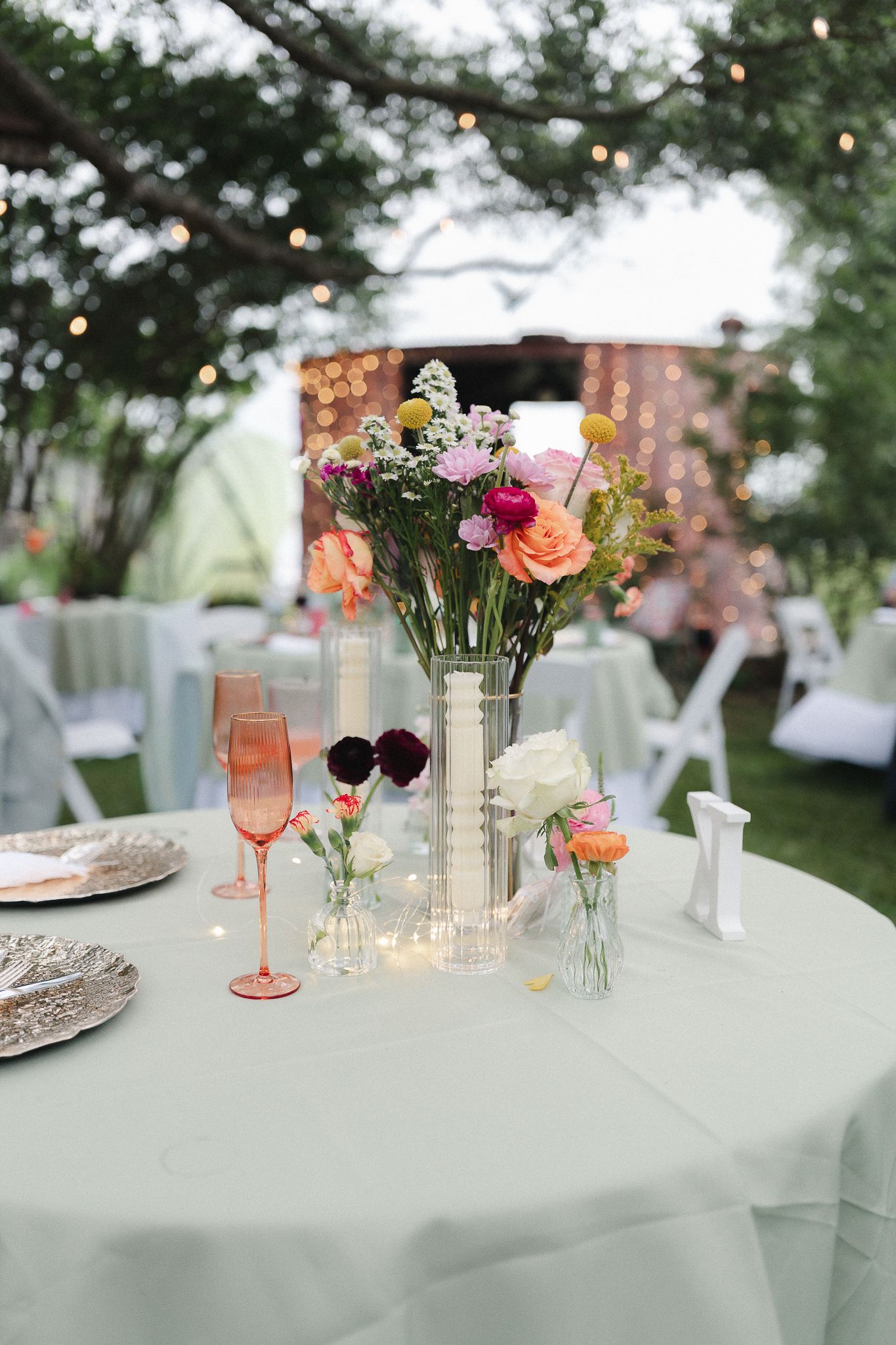 Outdoor wedding reception table with colorful floral centerpiece, pink champagne glasses, and string lights in background.