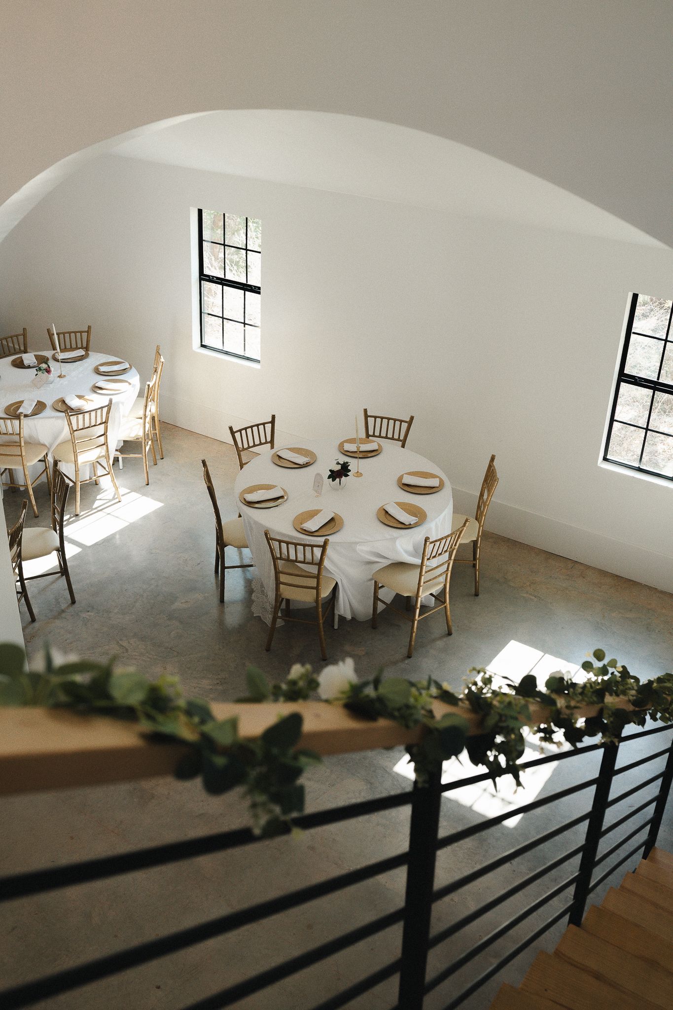 Elegant wedding reception venue with round tables, gold Chiavari chairs, white linens, and greenery-adorned staircase railing.
