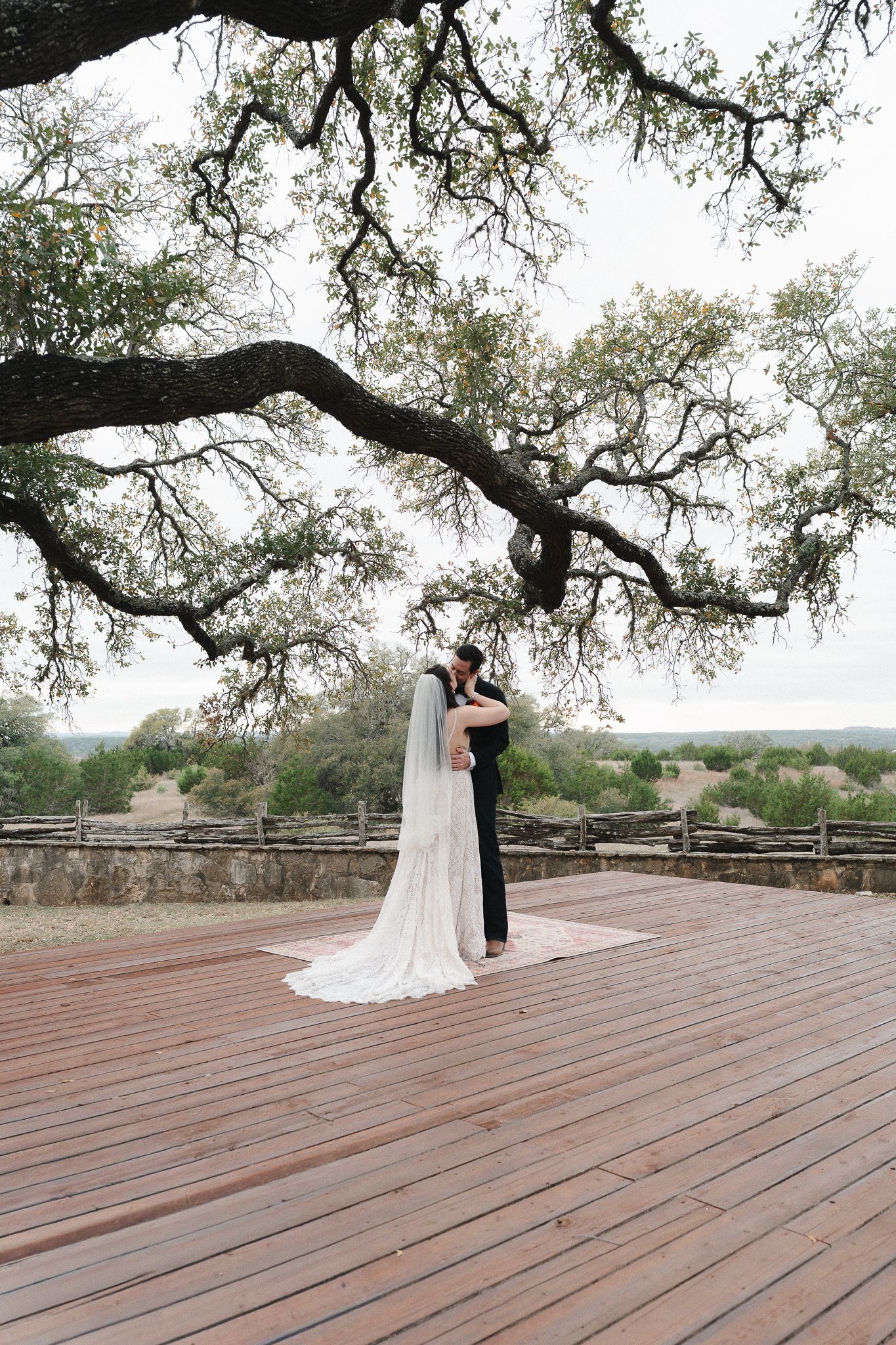 Bride and groom embracing on a wooden deck under a sprawling oak tree at an outdoor Texas Hill Country wedding.