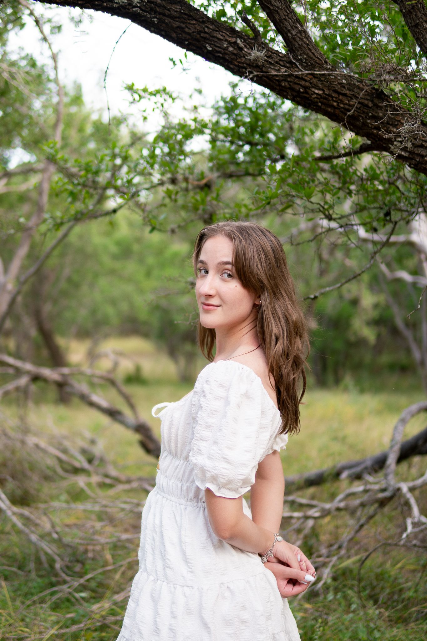 A person in a white lace dress poses gracefully among blooming spring trees in a natural outdoor setting.