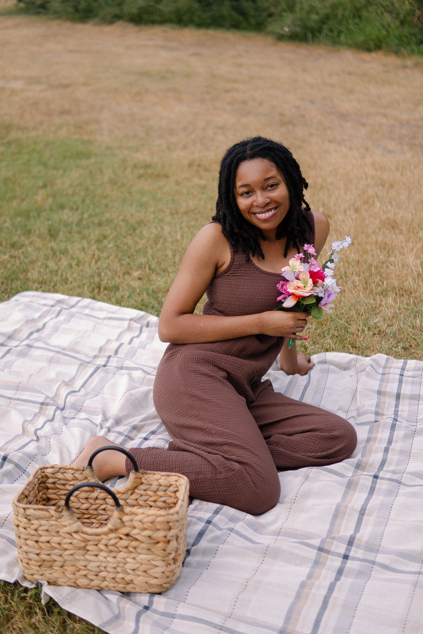 A person in a brown jumpsuit sits on a picnic blanket holding flowers with a woven basket nearby in a grassy field.
