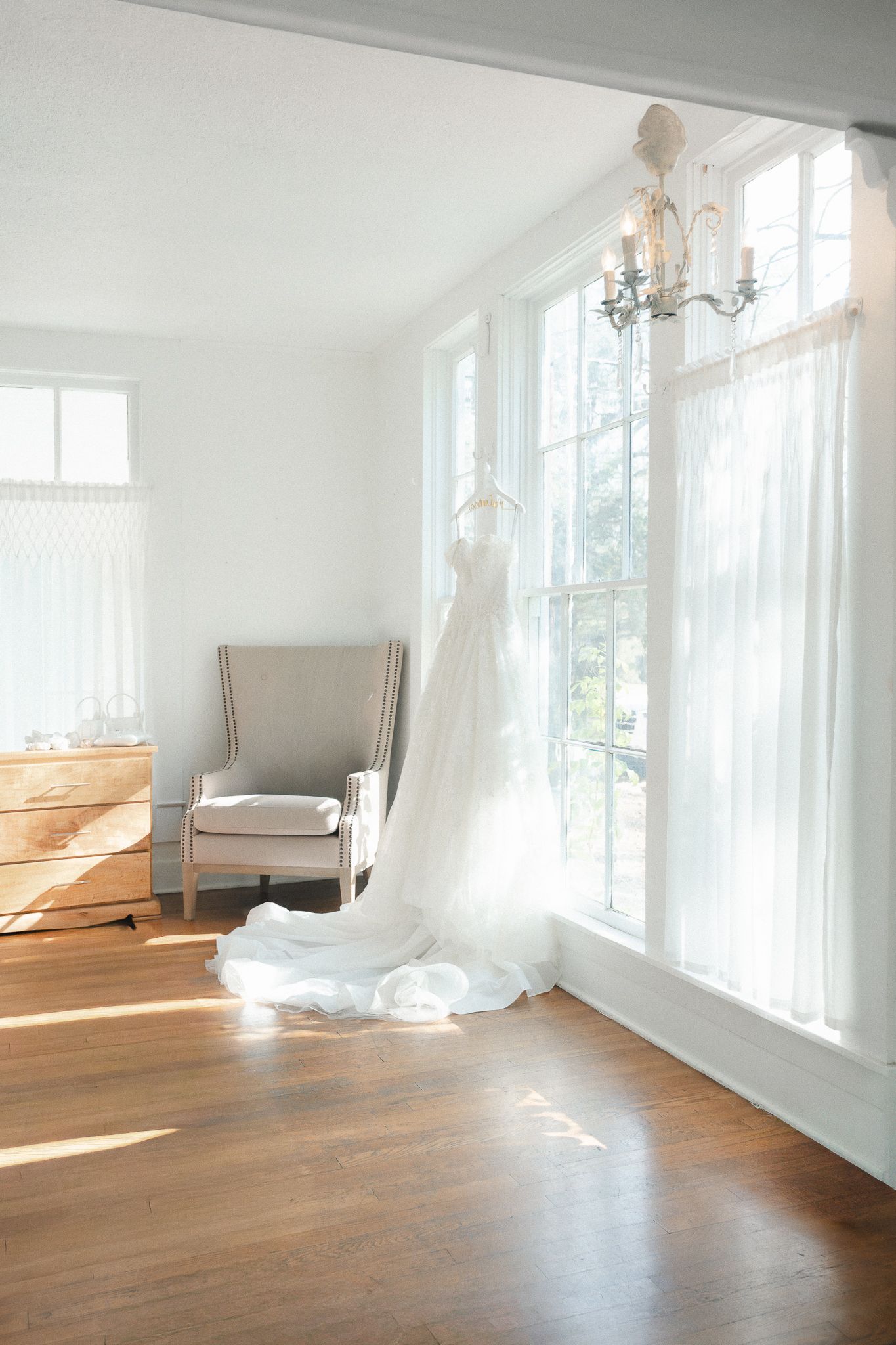 Elegant white wedding dress hanging by sunlit windows in a bright, airy bridal suite with hardwood floors.