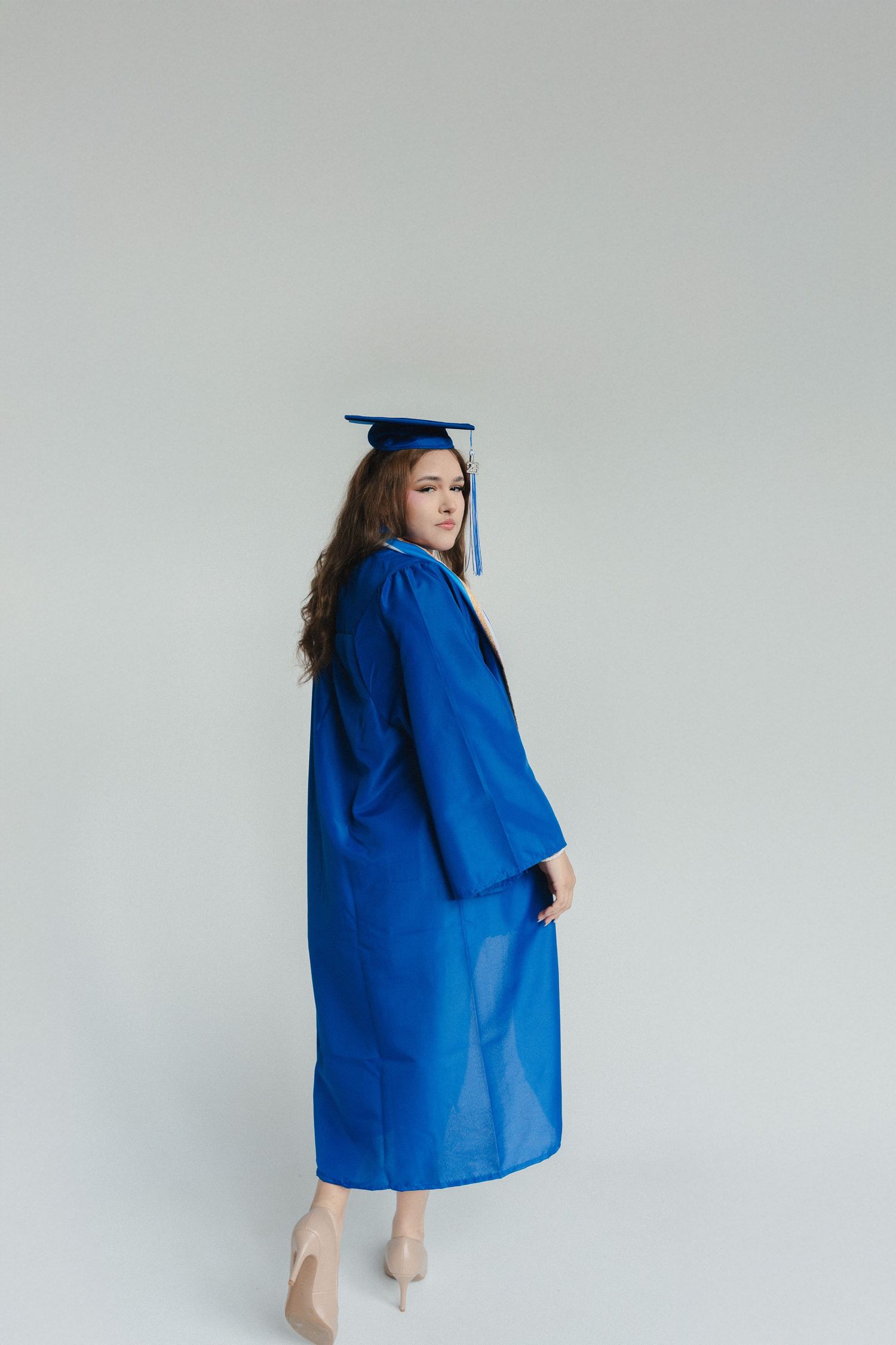 Young woman in blue graduation cap and gown posing in studio, looking over shoulder.