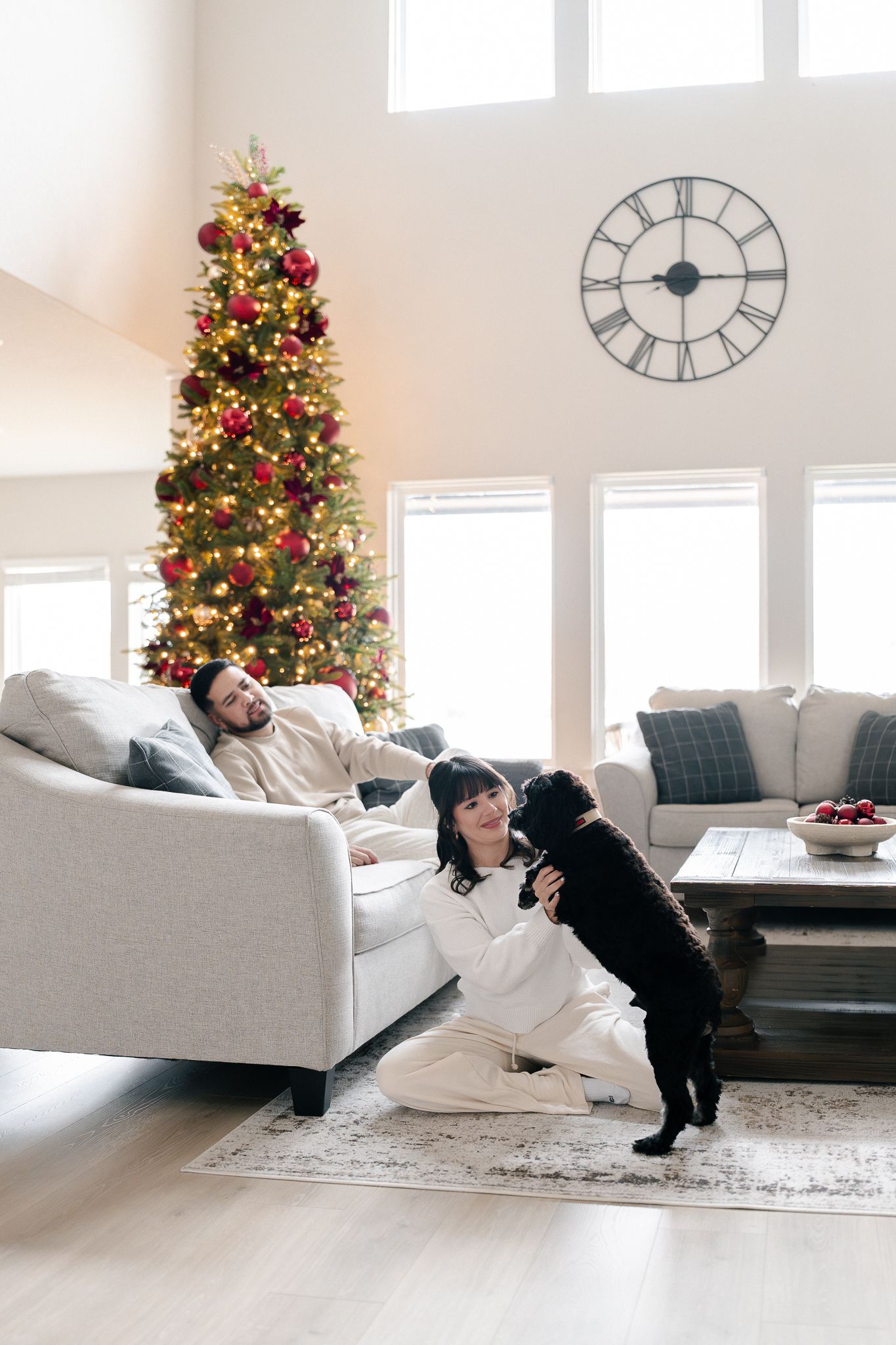 A cozy living room scene with a decorated Christmas tree and a playful black dog interacting with people on a beige couch.