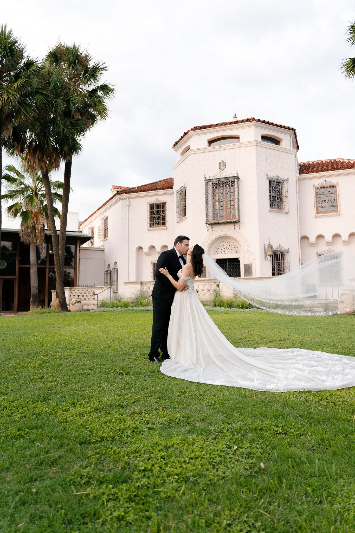 A couple embraces on a lawn in front of a Spanish-style white building with red tile roof and palm trees.