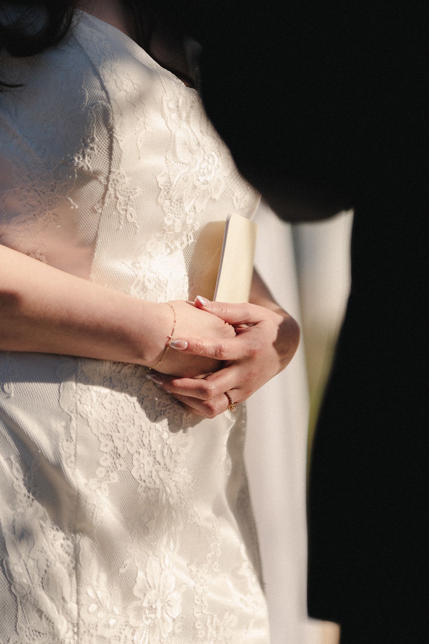 Bride in white lace wedding dress holding a small book during ceremony, wearing delicate gold bracelet and ring.