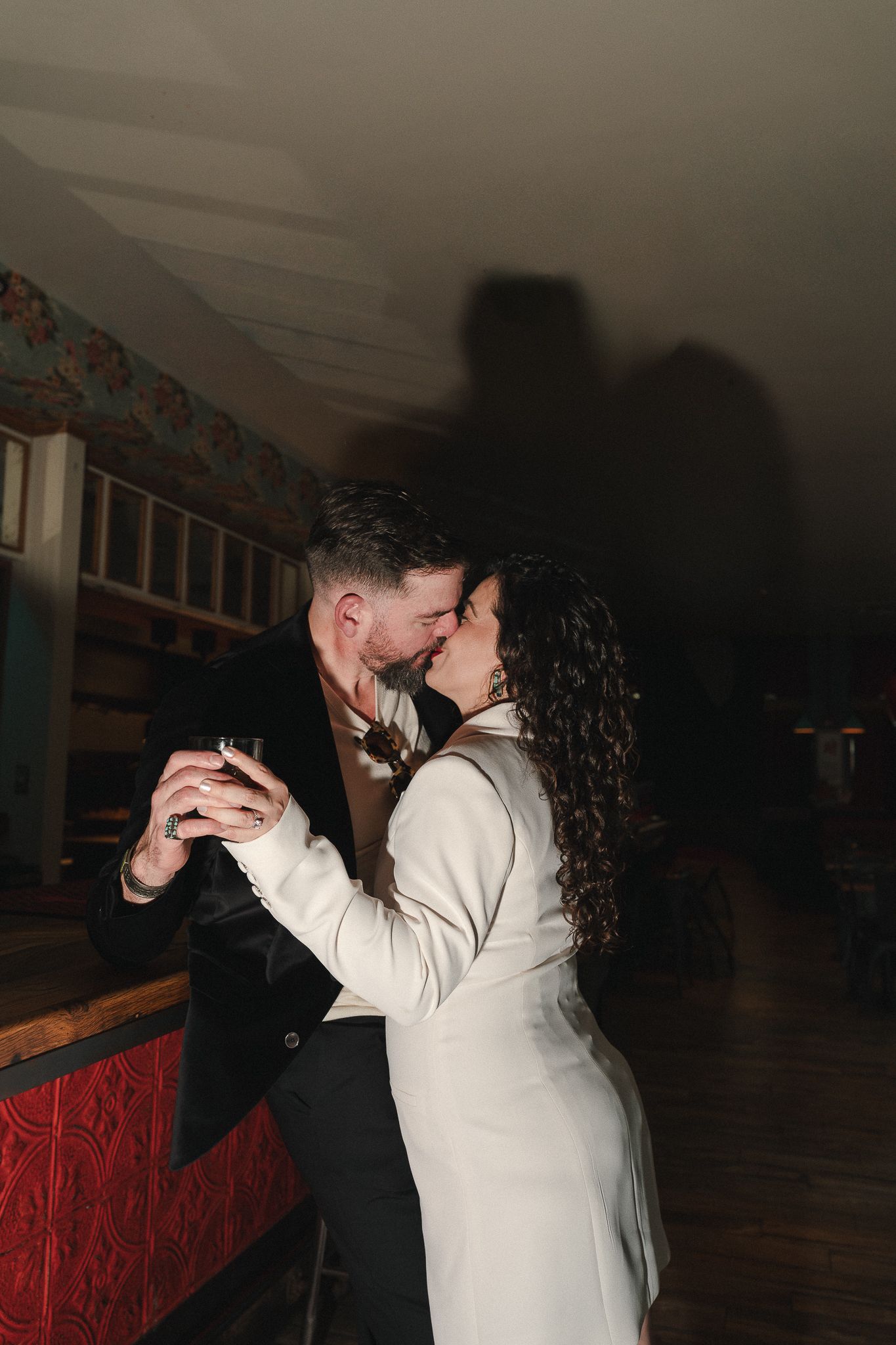 Couple sharing a romantic kiss while holding drinks in a dimly lit bar, woman in white blazer, man in black suit.