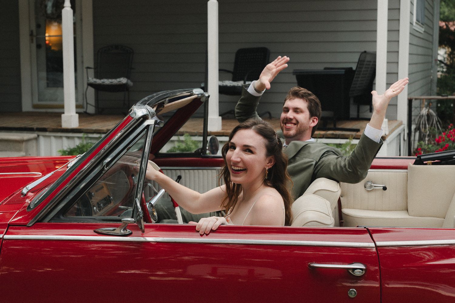 Happy couple in wedding attire celebrating in a classic red convertible car outside a house.