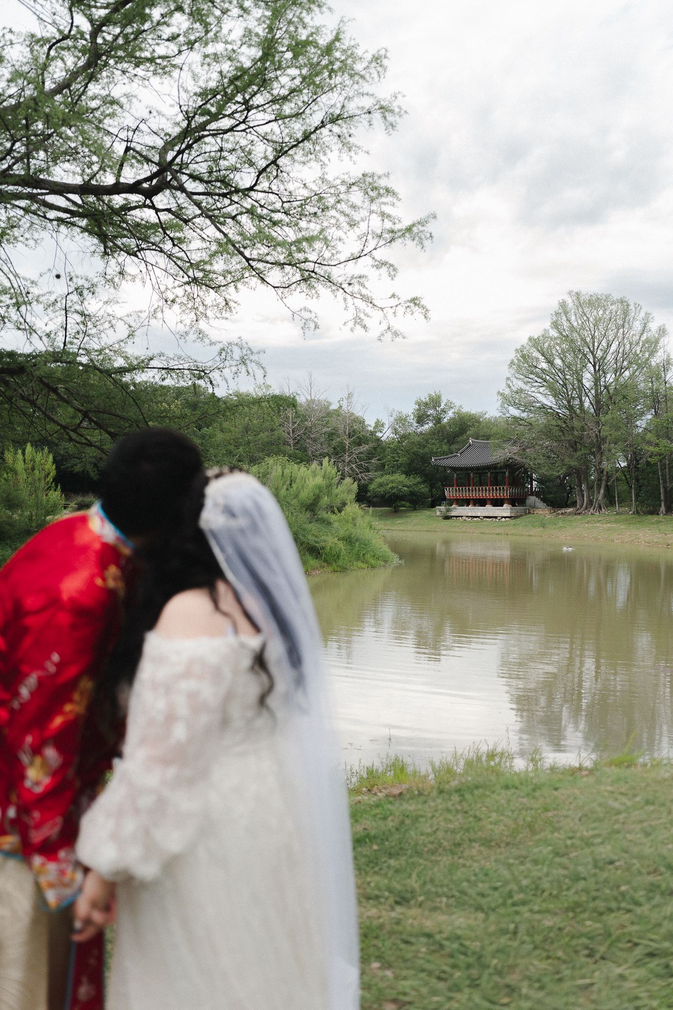Bride in white dress and groom in red traditional attire stand by a serene pond with a pavilion in background.