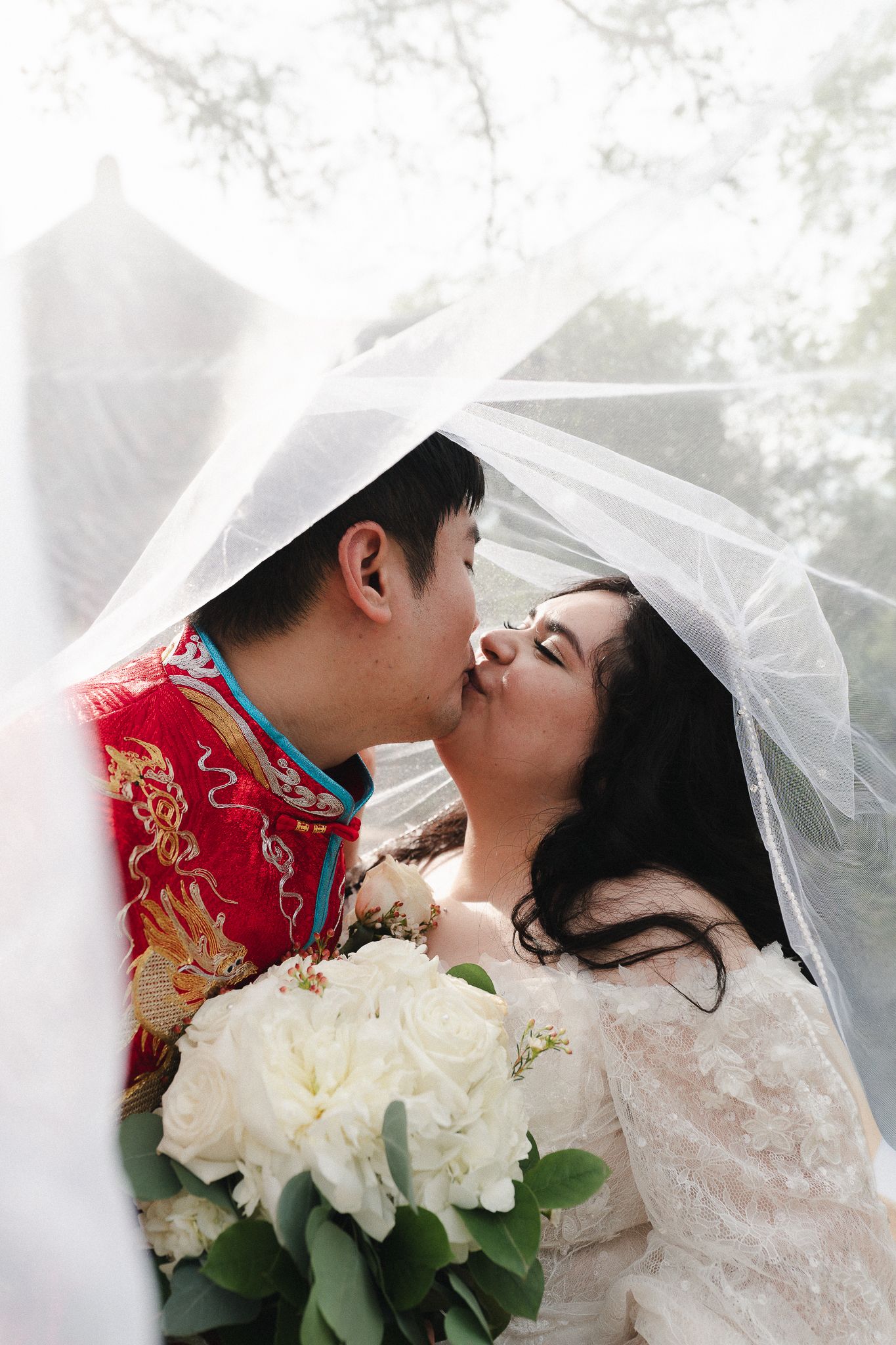 Bride and groom sharing a kiss under a white veil, groom in red Chinese wedding attire, bride holding white peony bouquet.