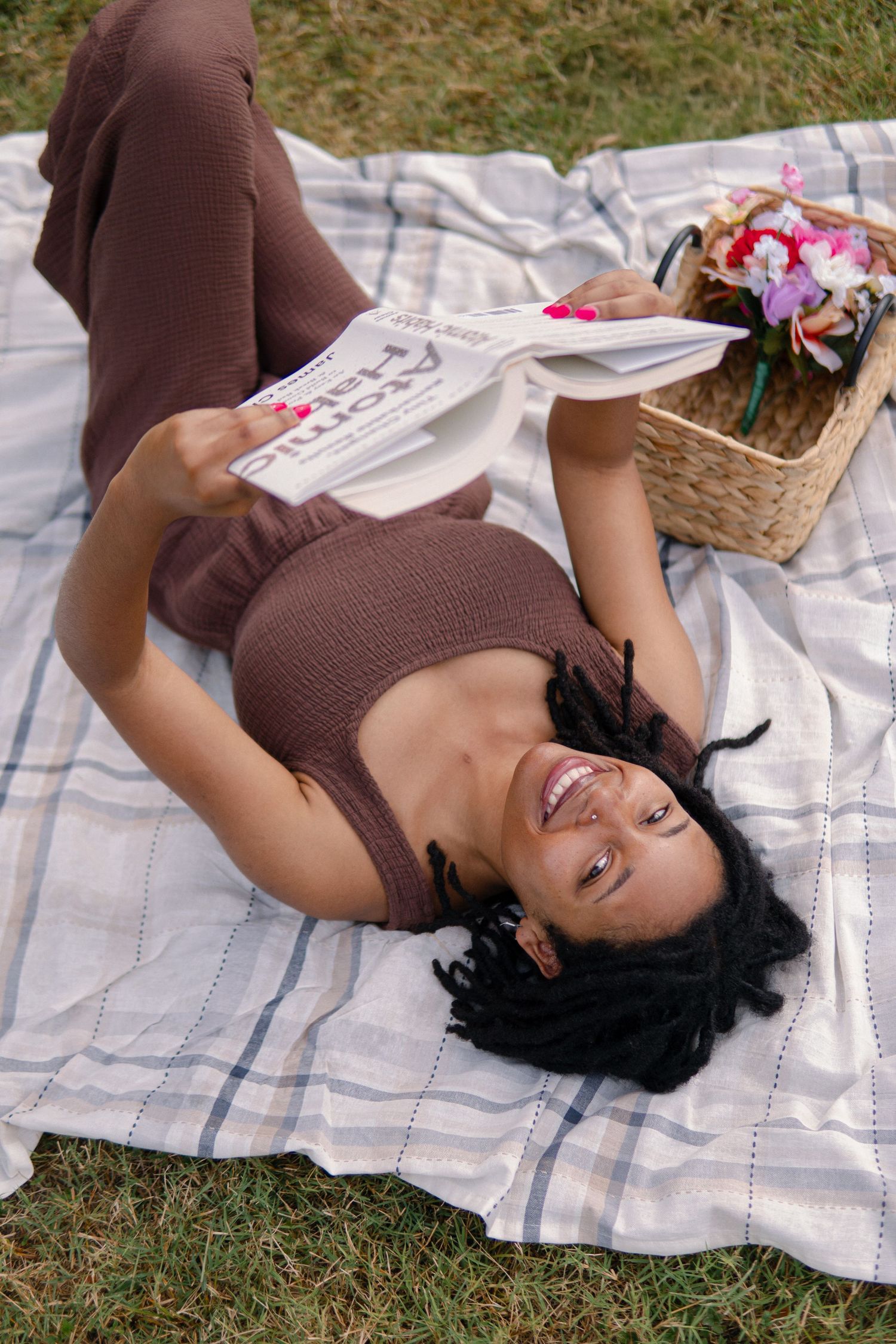 Someone relaxes on a picnic blanket while reading a book next to a wicker basket with flowers on a sunny day.