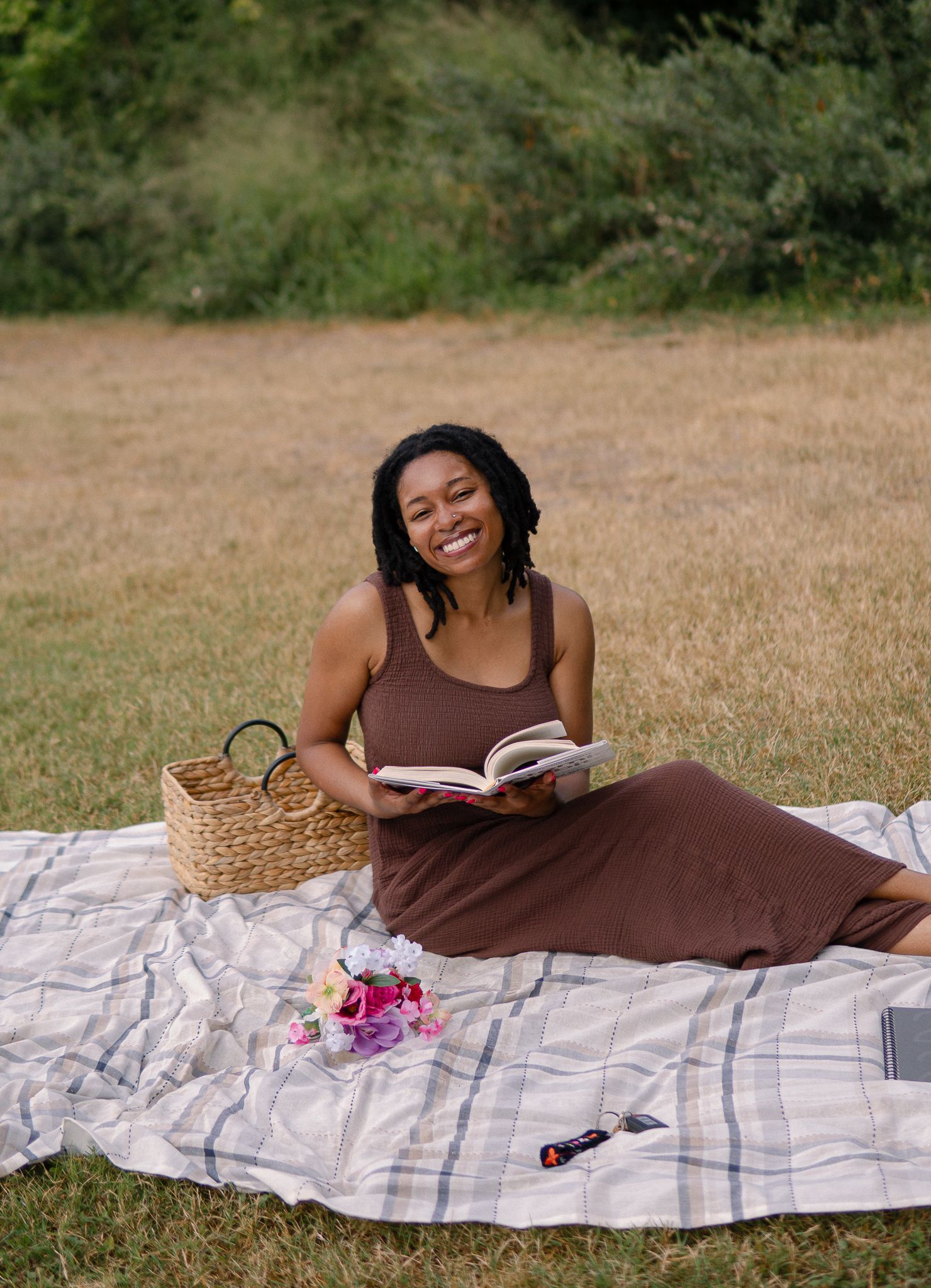Someone wearing a brown dress relaxes on a plaid blanket while reading a book outdoors on a sunny day.