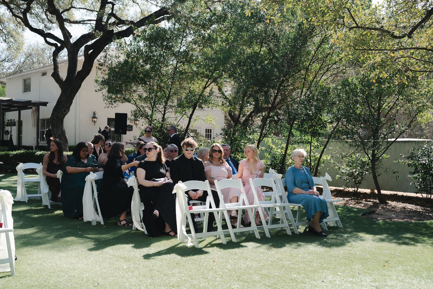 Guests seated in white chairs at an outdoor wedding ceremony on green lawn surrounded by trees.