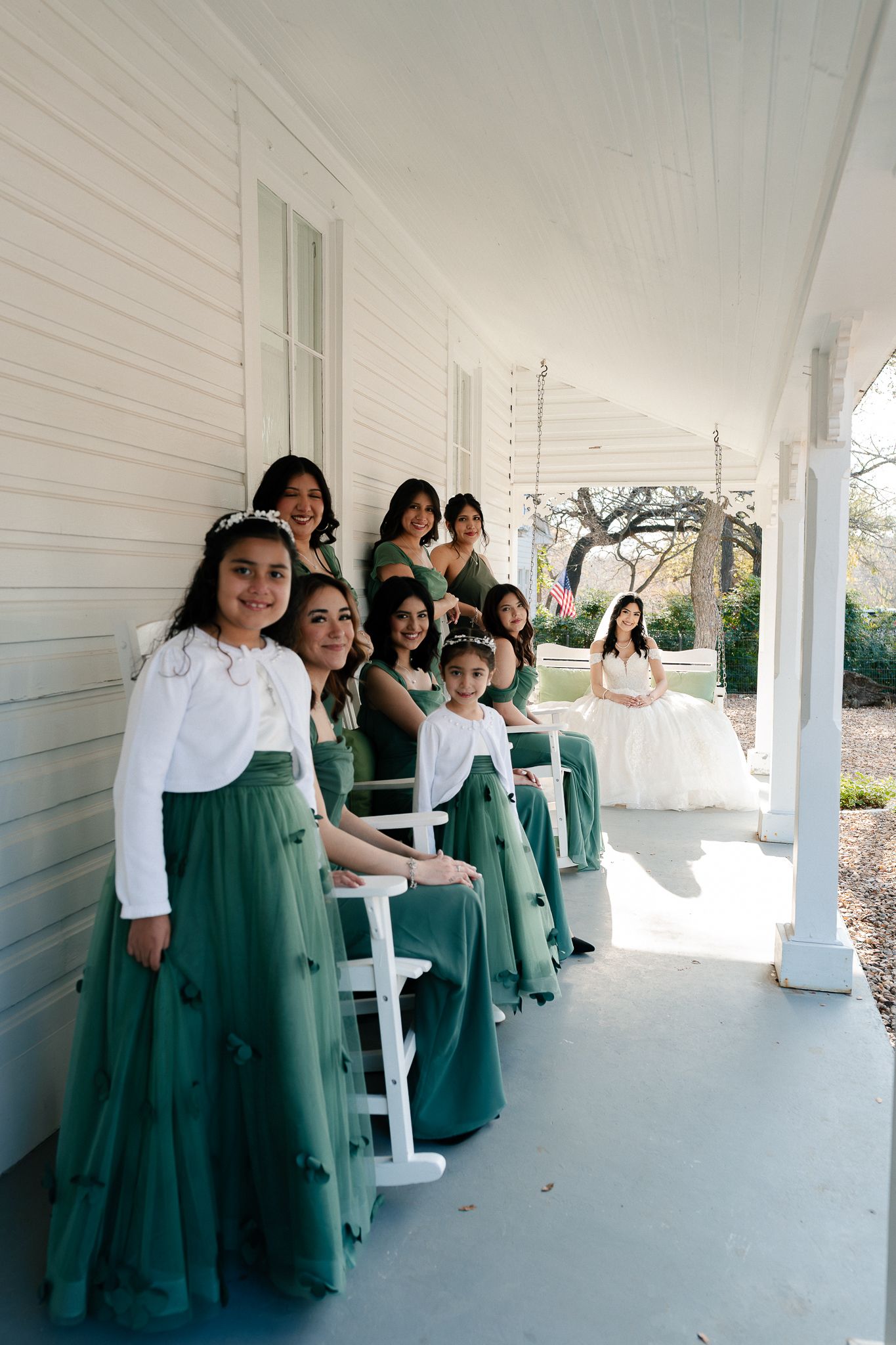 Bridesmaids in matching emerald green dresses pose together on a white wraparound porch.