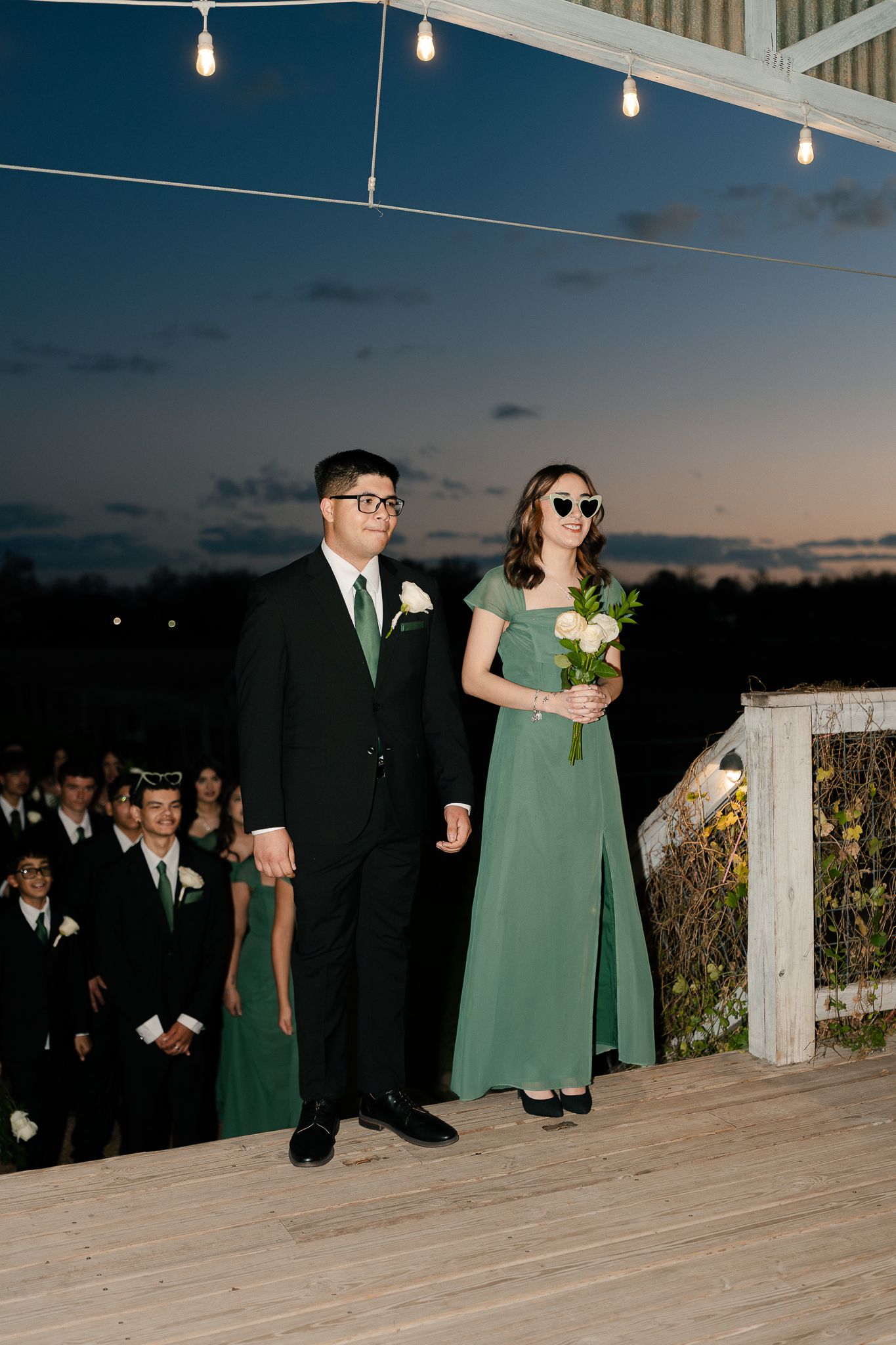 Wedding party members in coordinated green and black attire stand on a wooden deck at sunset under string lights.