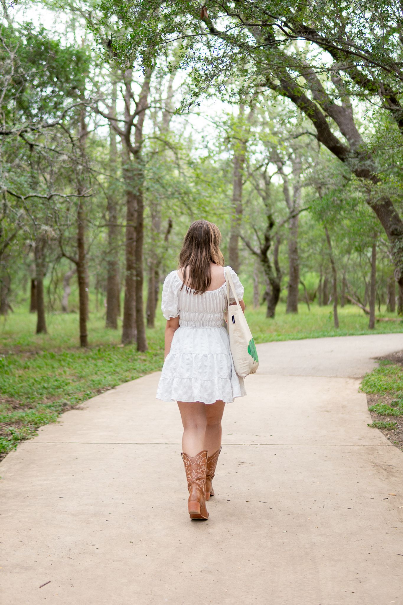 A person in a white top and brown pants walks away on a winding dirt path through a green wooded area.