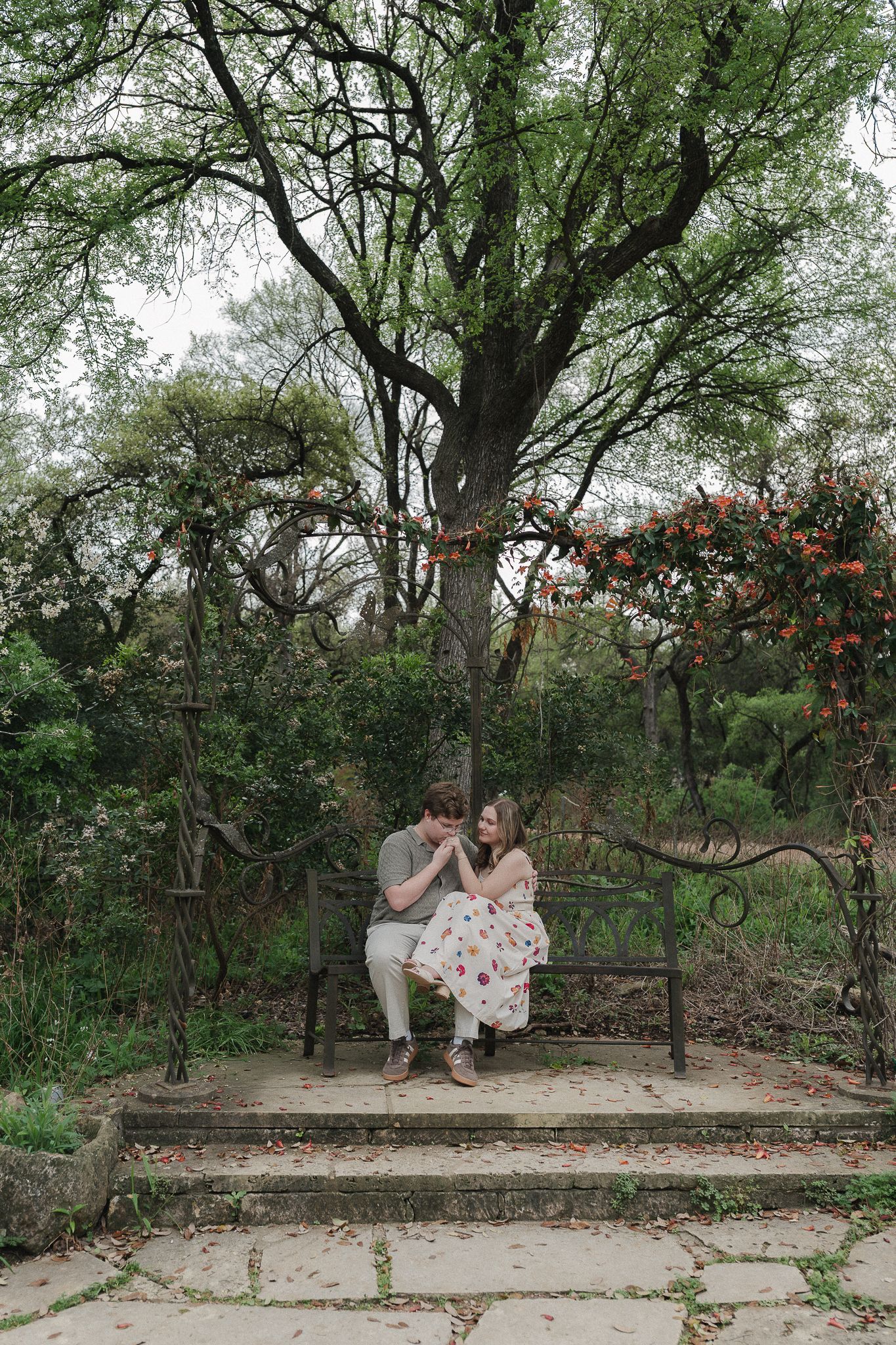 A couple sits closely on a wrought iron bench in a lush garden, surrounded by green trees and flowering vines.