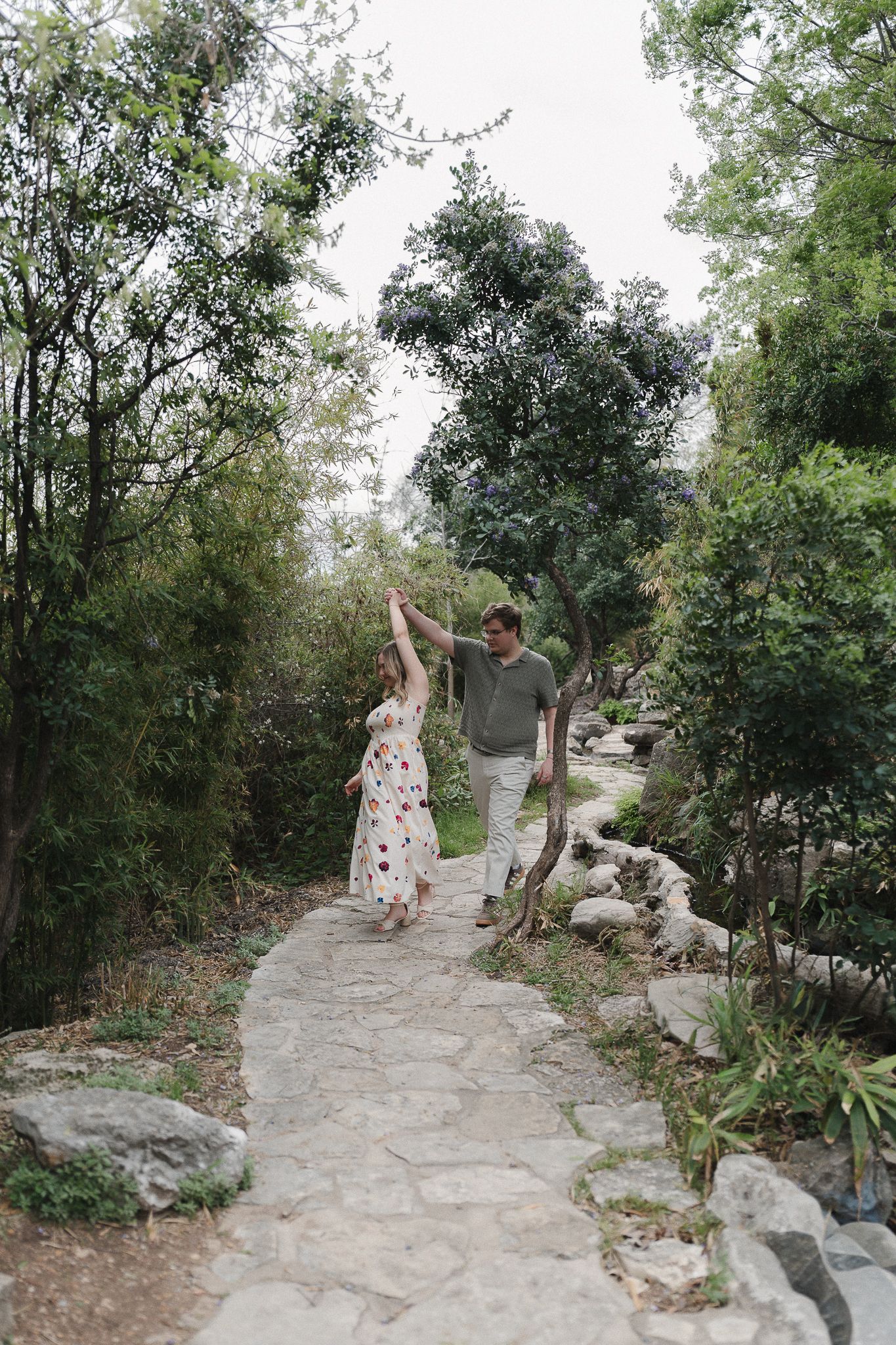 Couple holding hands and dancing on a stone garden path surrounded by lush green trees and plants.