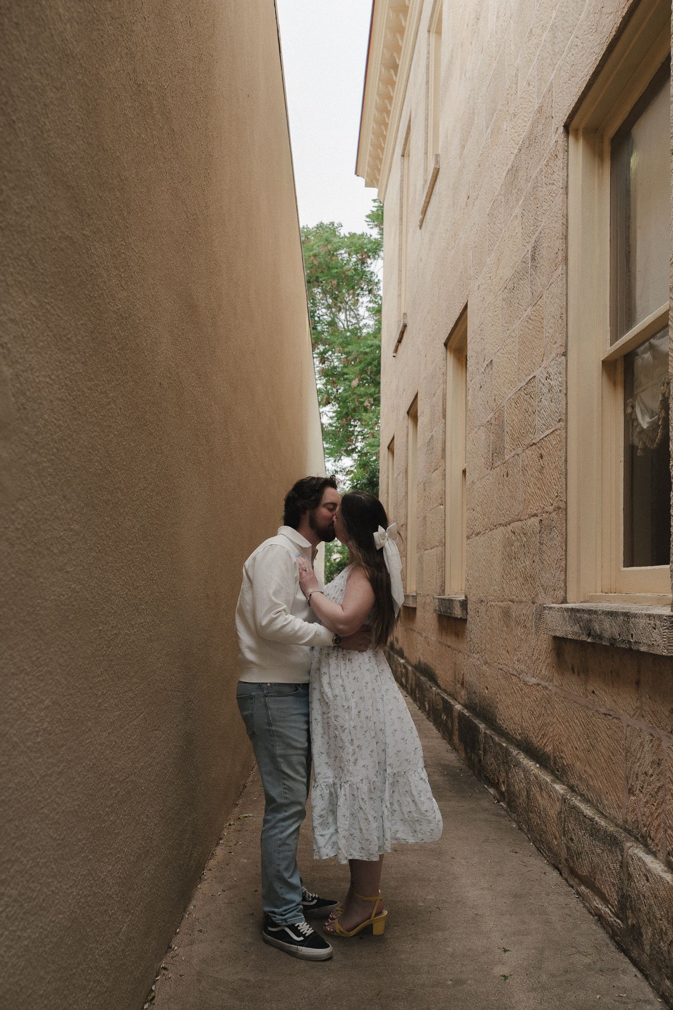 Couple sharing a romantic kiss in a narrow alleyway between two stone buildings, woman in white dress, man in jeans.