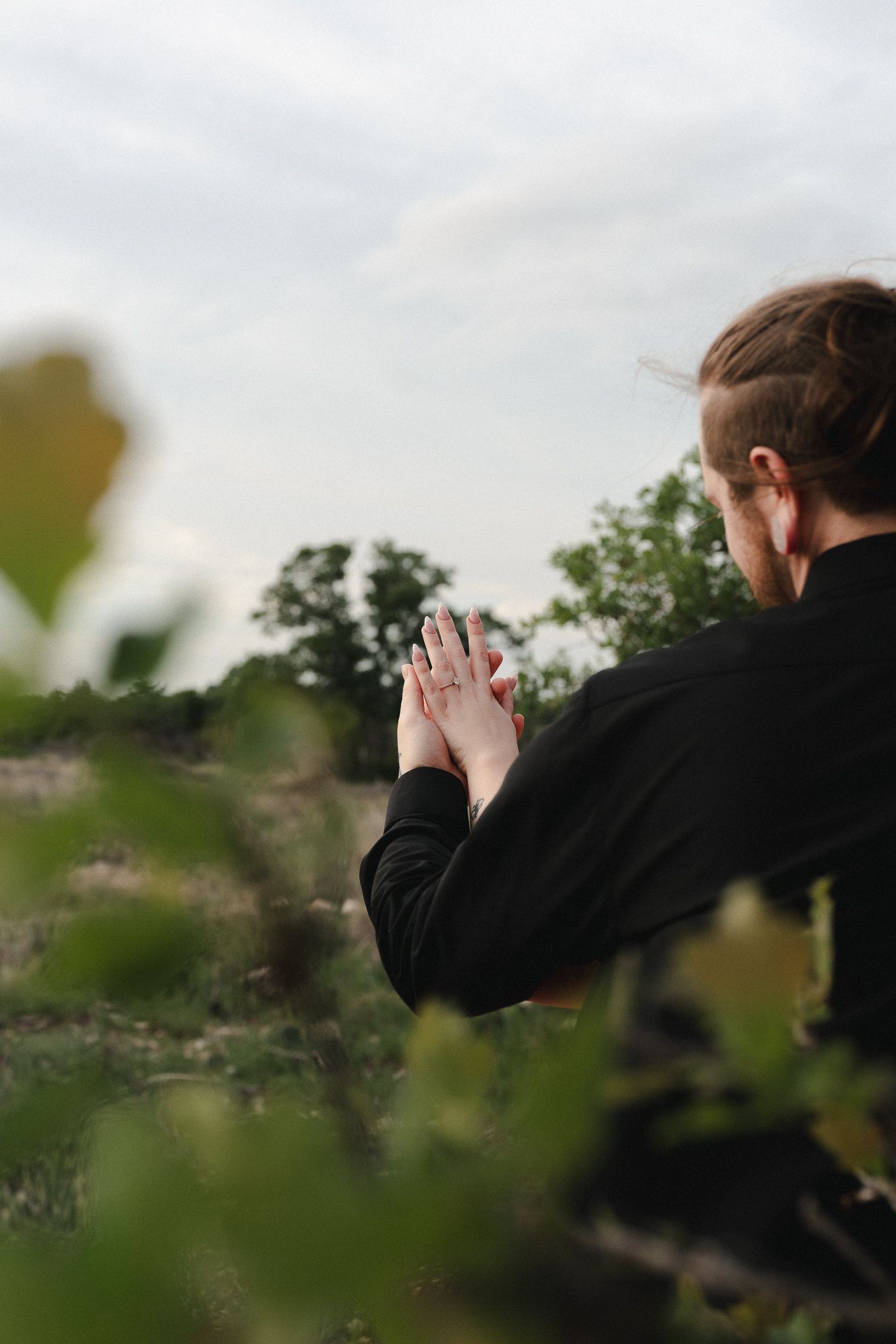 Man in black jacket holds hands with someone outdoors, surrounded by lush greenery under a cloudy sky.
