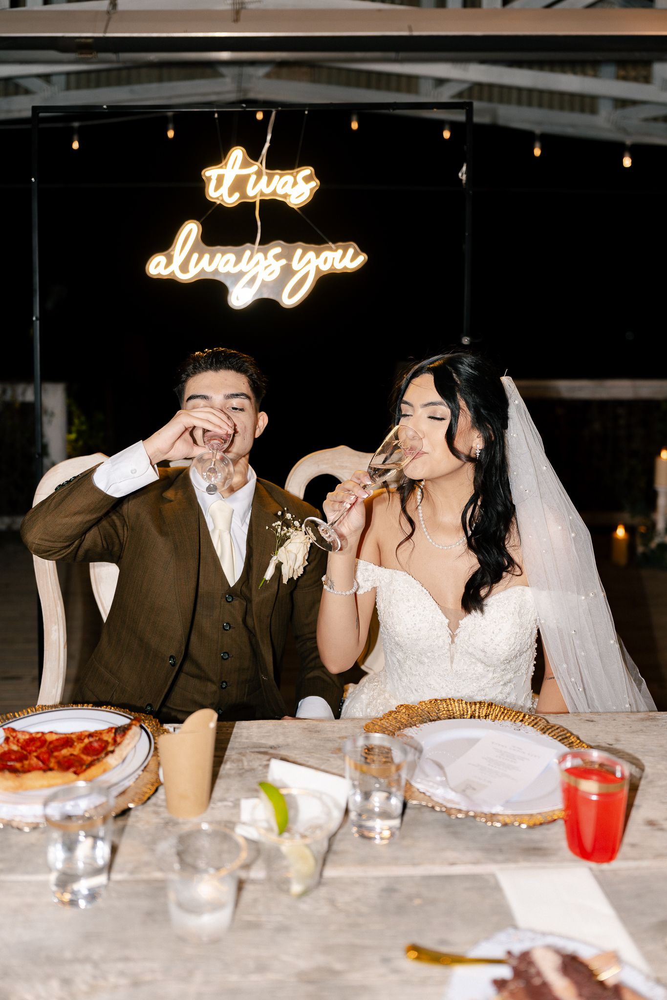 Newlyweds share a playful moment eating pizza at their reception under an illuminated 'always you' neon sign.