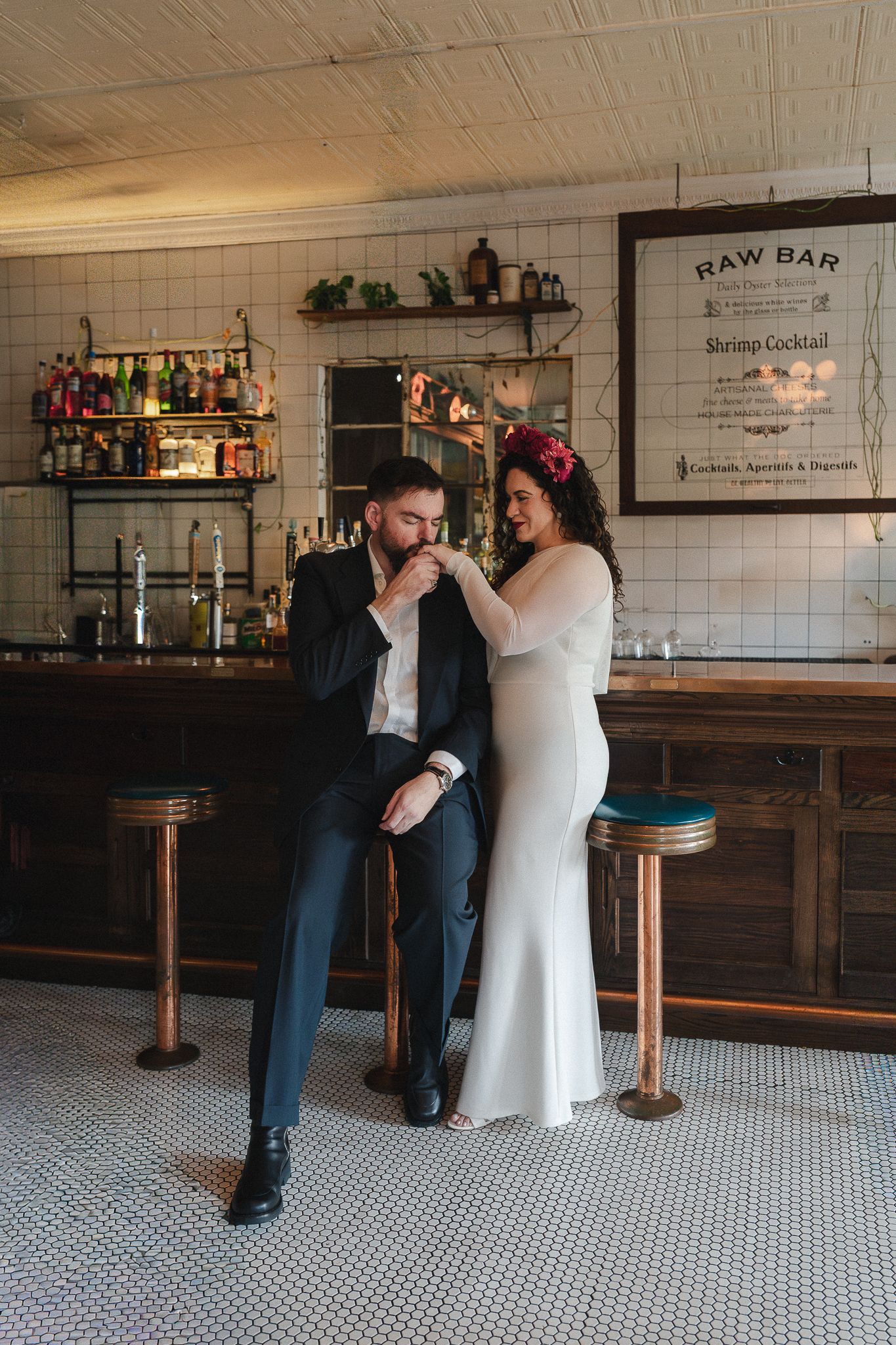 A groom kisses his bride's hand at a Raw Bar venue, she wears a white gown with floral hair piece.