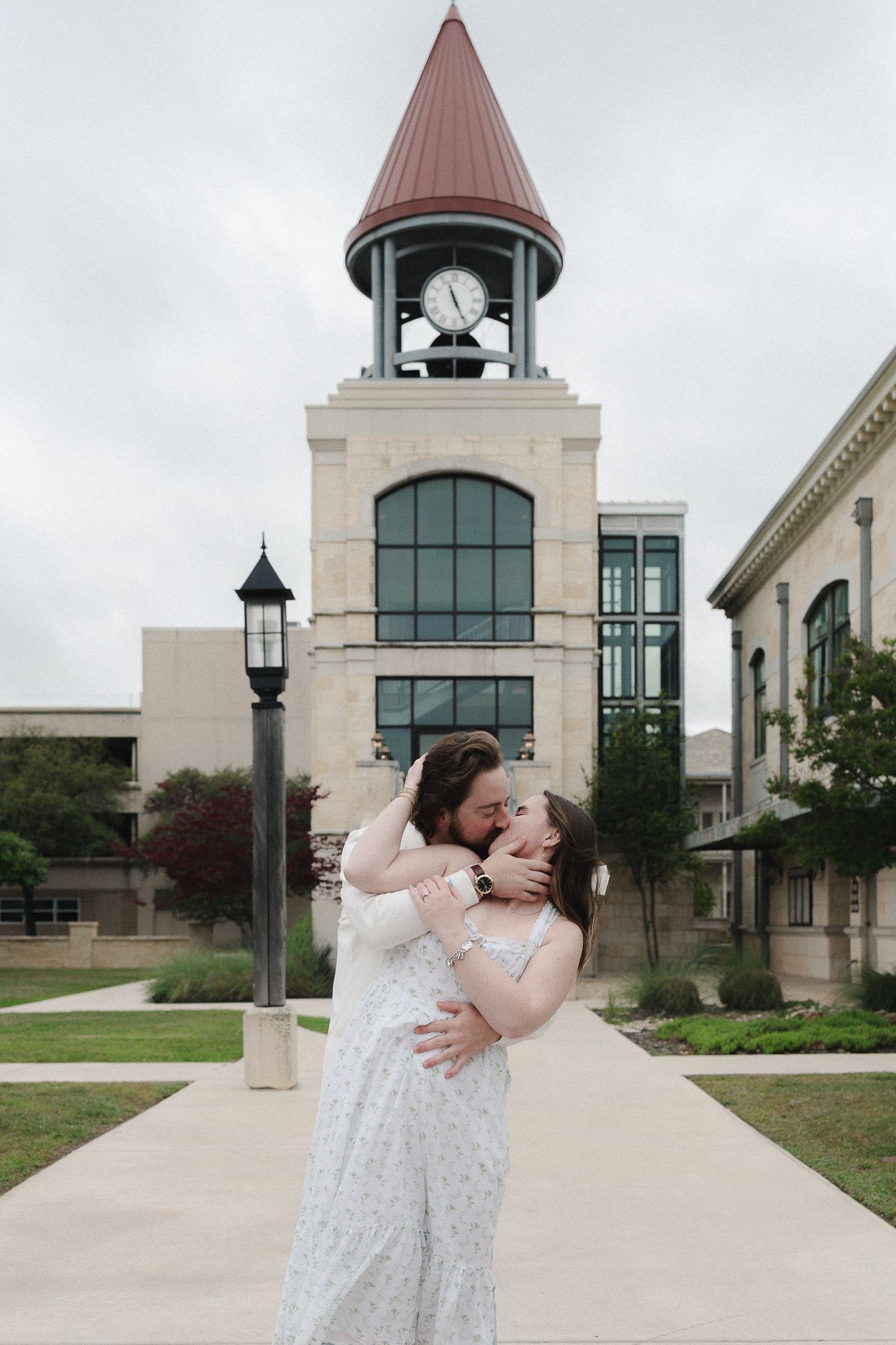 Couple embracing and kissing in front of a clock tower building on a cloudy day.