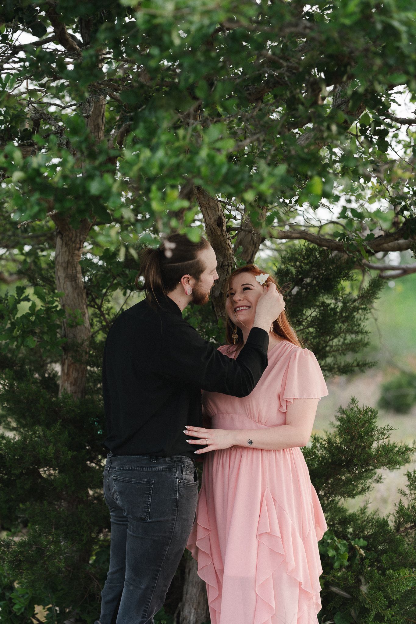 A couple shares a tender moment under a lush green tree, man in black touches womans red hair as she smiles in pink dress.