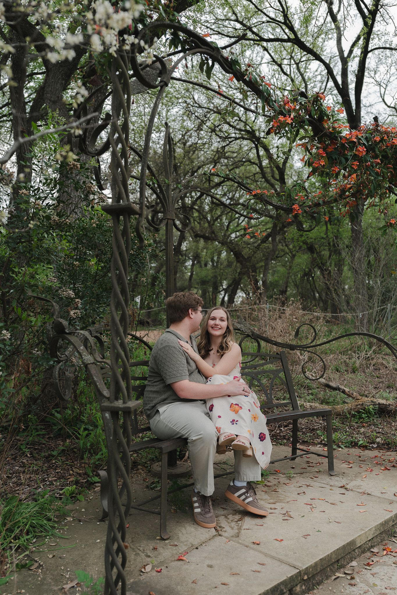 Couple sitting on ornate iron bench under floral arbor in lush garden, sharing a romantic moment.