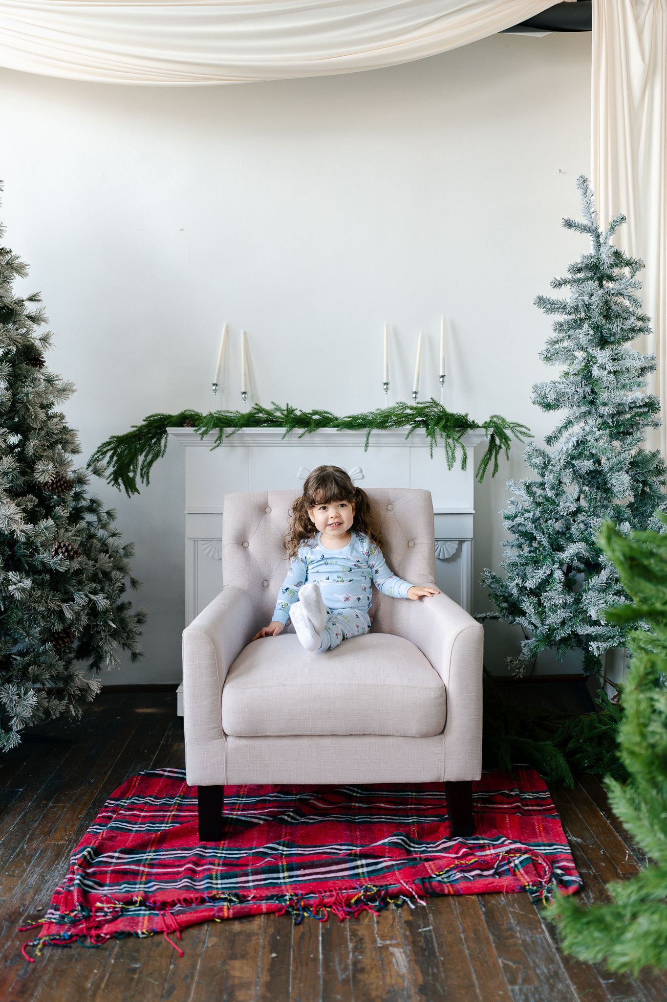 Holiday scene with armchair, plaid blanket, white mantel with greenery, and Christmas trees against white backdrop.