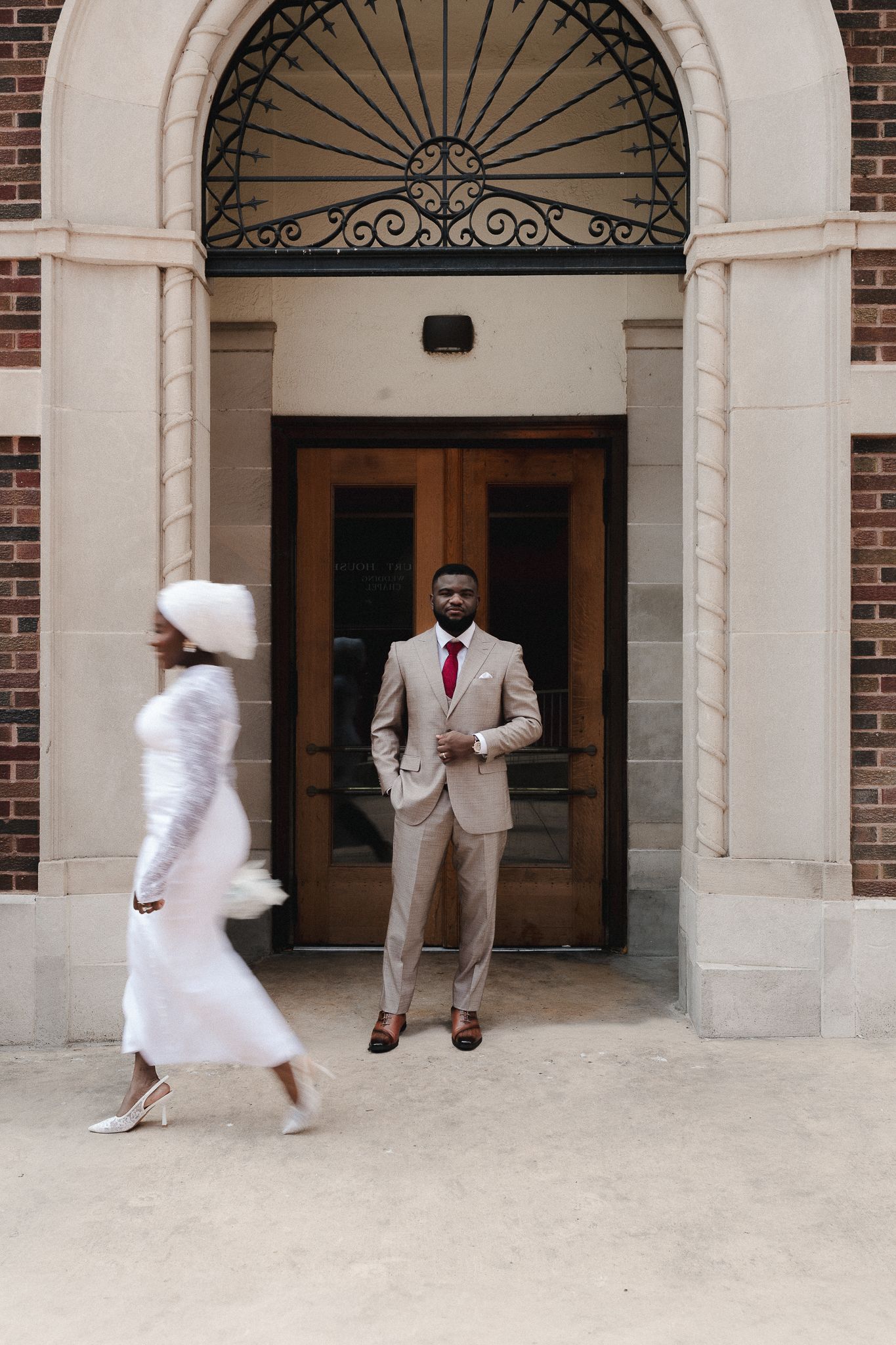 Groom in tan suit stands at ornate building entrance as bride in white dress and fur stole walks past.